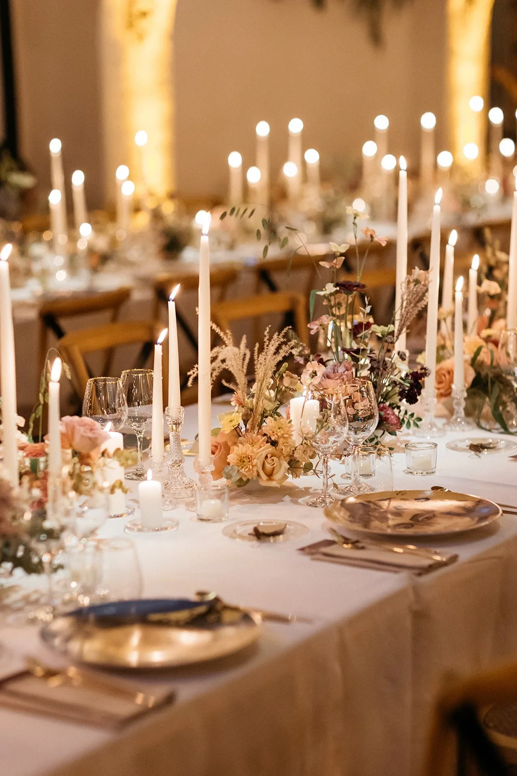 Elegant banquet table decorated with tall white candles, floral arrangements, wine glasses, and gold-rimmed plates, set for a formal event or wedding reception.