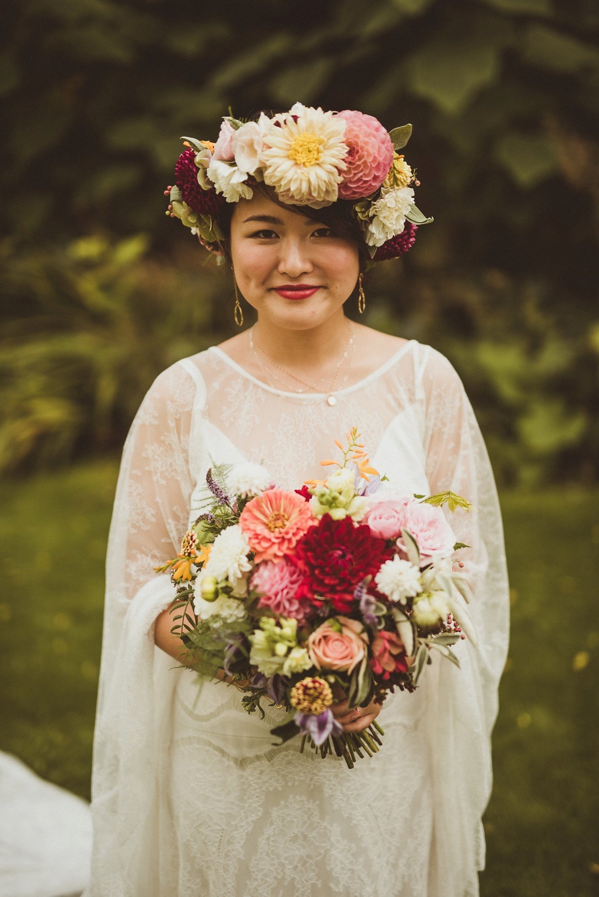 A woman wearing a lace dress and a floral crown made of large, colorful flowers holds a bouquet of various flowers. She has short dark hair, gold earrings, and is smiling against a green, blurred outdoor background.
