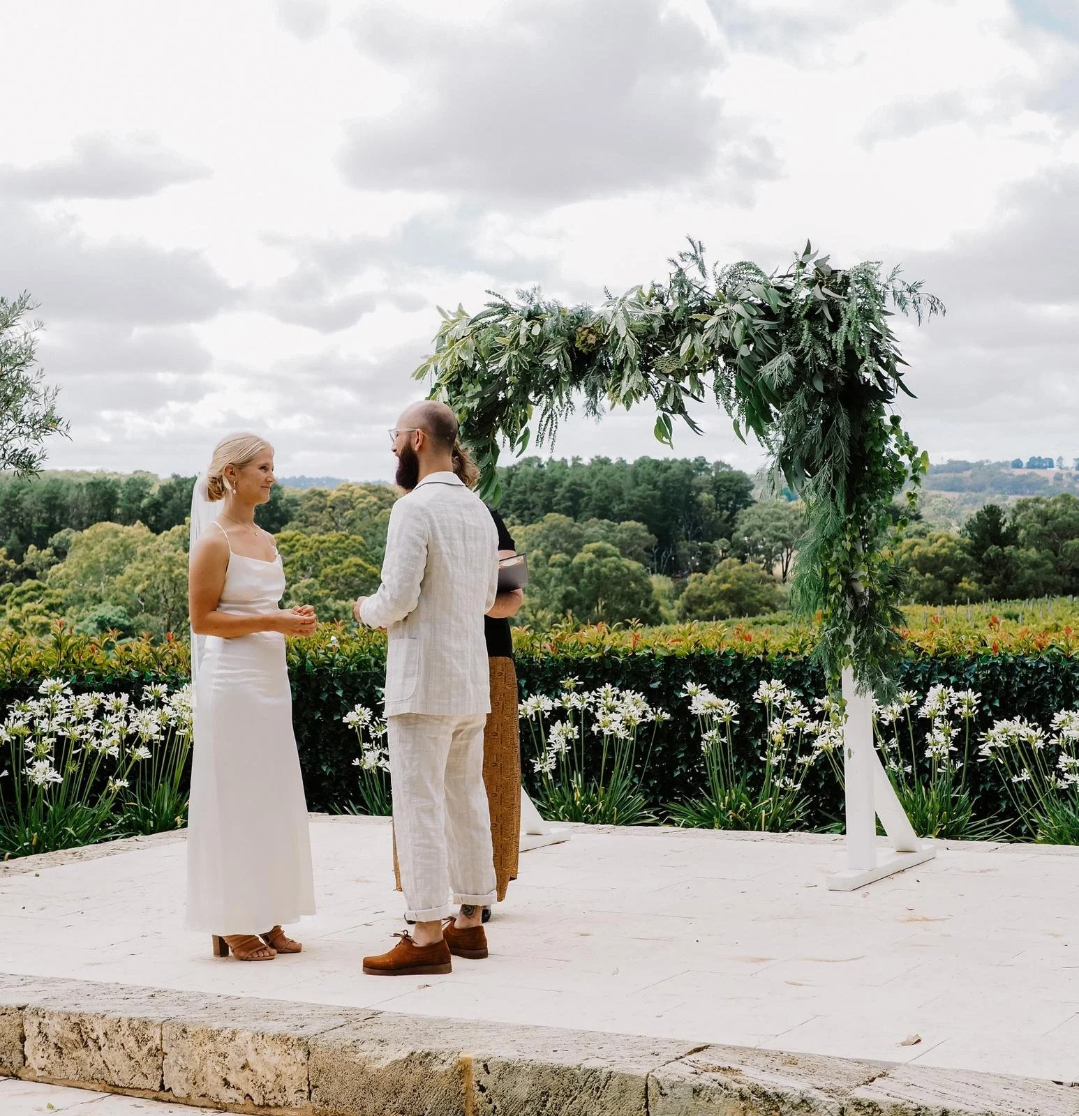 A couple getting married outdoors during the day, with a woman in a white strapless dress and a man in a cream-colored suit holding hands in front of an officiant. The ceremony is under a green leafy arch, with a scenic landscape of trees and hills i