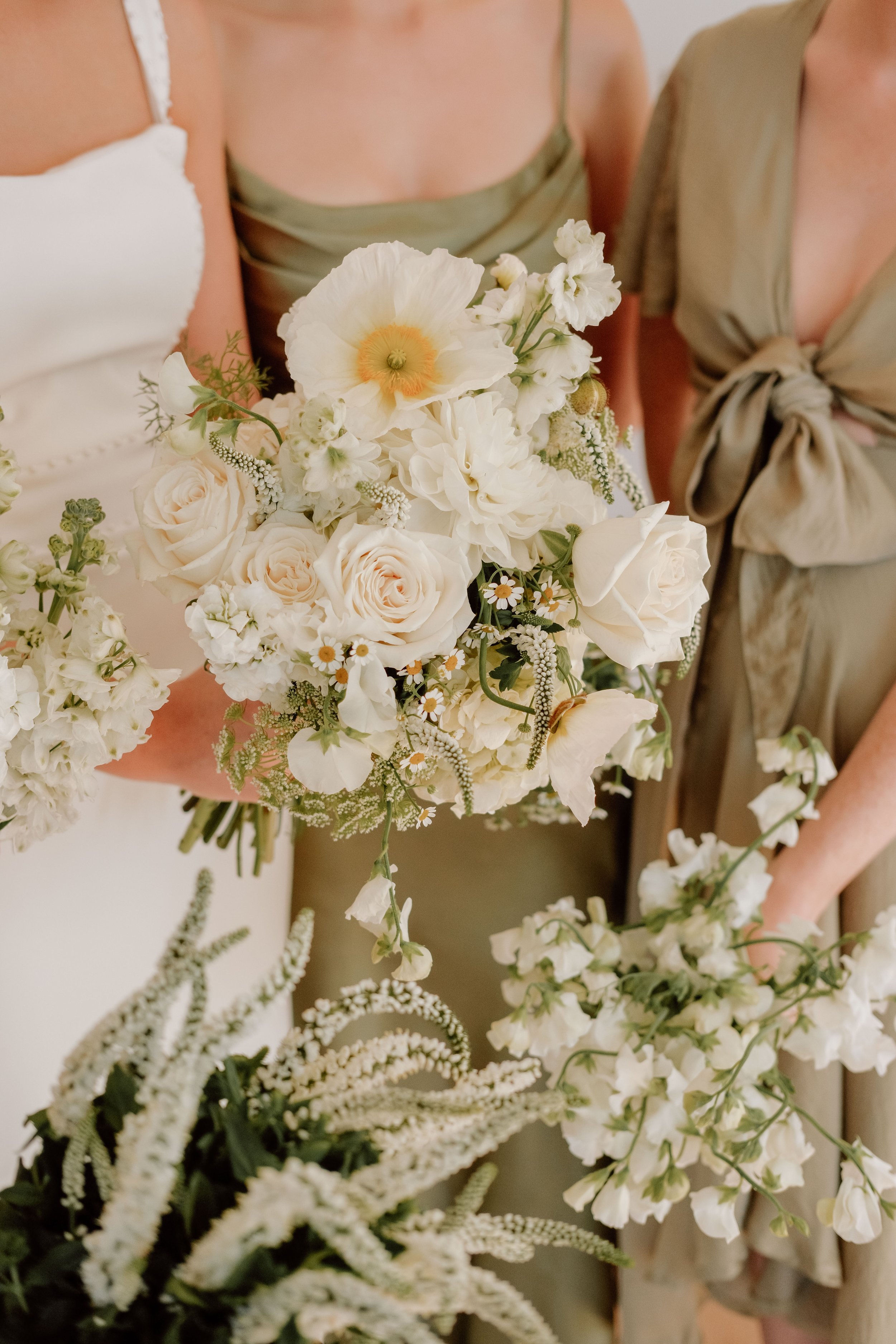 Close-up of three women in dresses holding a bouquet of white and cream flowers, including roses, lilies, and daisies, at a celebration or wedding.
