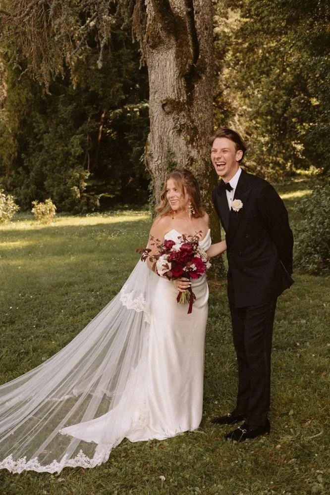 A bride and groom standing outdoors in front of a large tree, smiling and laughing. The bride wears a white wedding dress with a long veil and holds a bouquet of dark red and pink flowers. The groom wears a black tuxedo with a white shirt and black b
