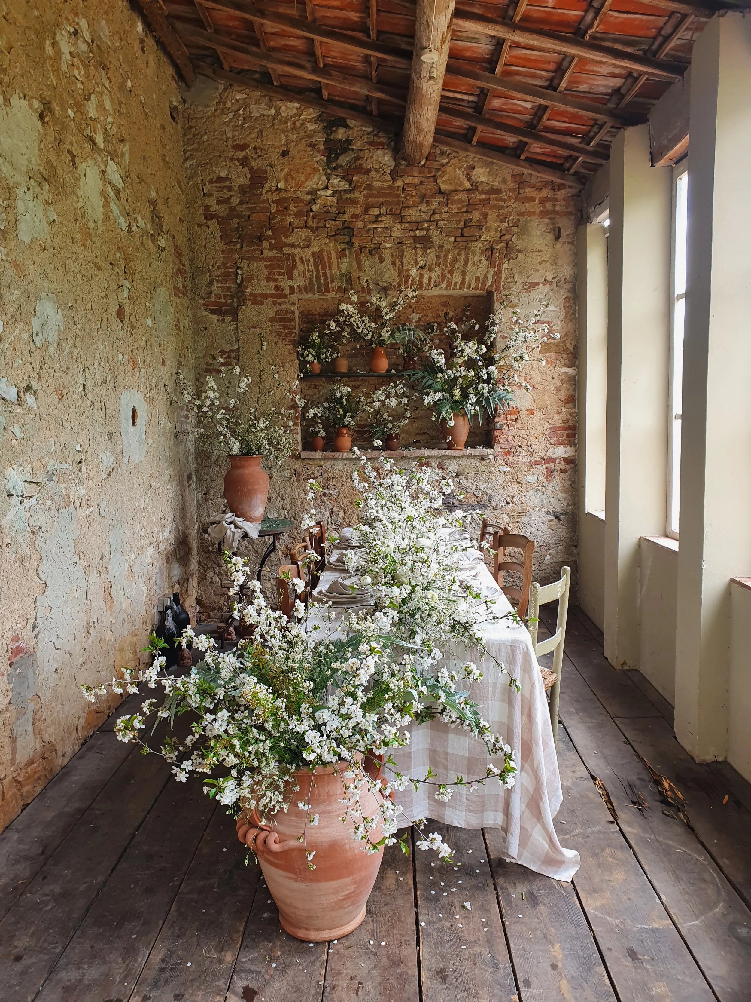 A rustic dining area with a long table covered with a white checkered tablecloth and decorated with a large arrangement of white flowers and greenery in a terracotta pot. The room has exposed brick walls, a wooden ceiling, and natural light coming th