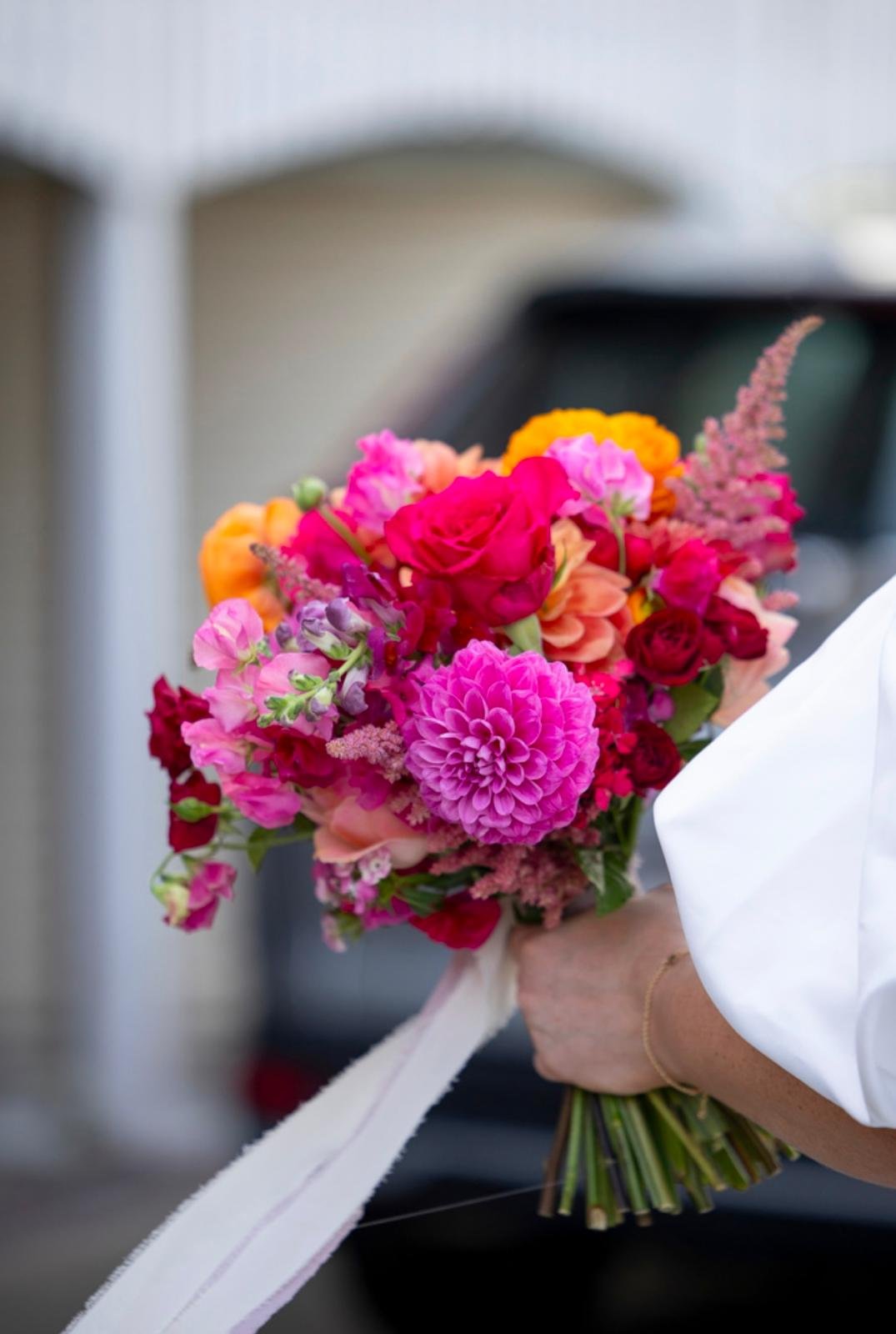 Person holding a colorful bouquet of mixed flowers including pink, red, orange, and purple blooms, with a white ribbon tied around the stems.