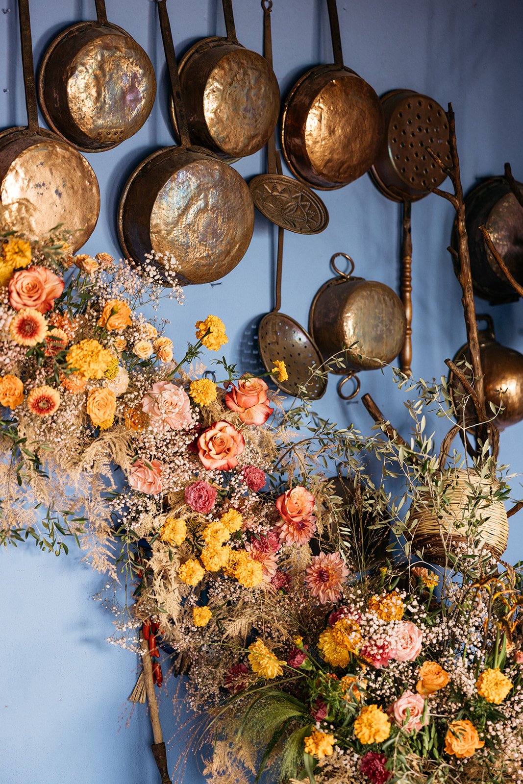 Group of vintage hanging copper cooking pots on a blue wall, with a colorful floral arrangement in the foreground.