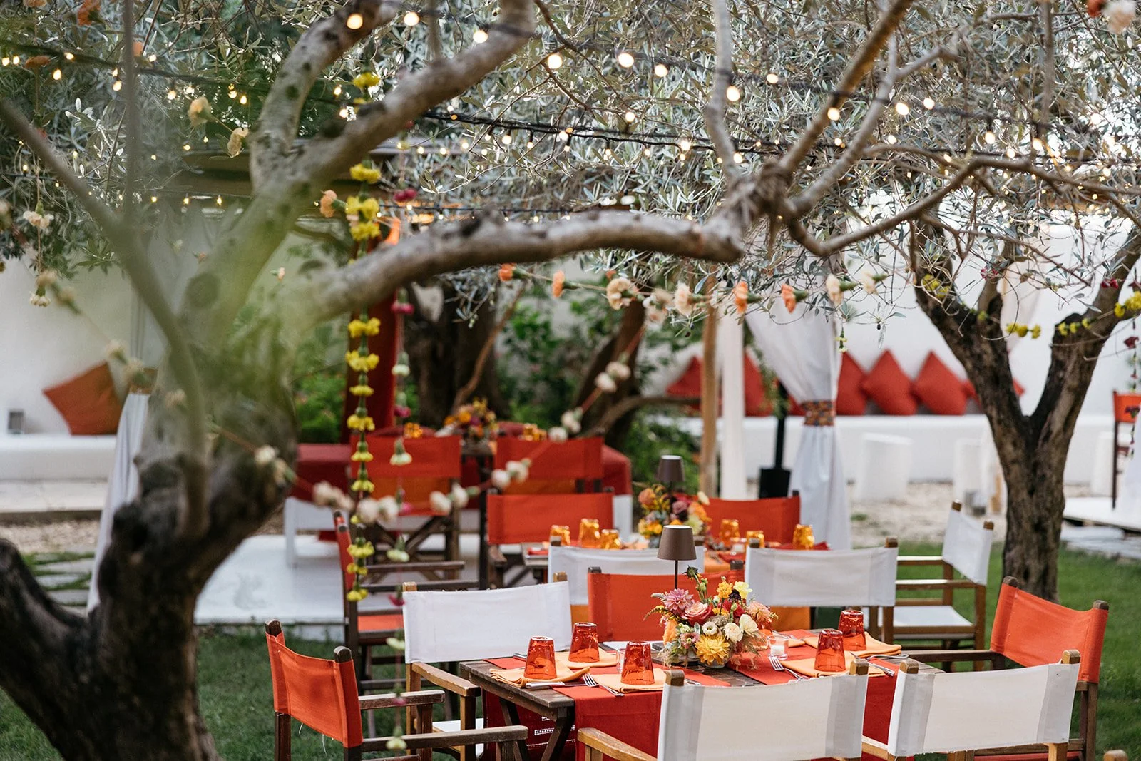 Outdoor dining setup with tables decorated with orange tablecloths and floral centerpieces, surrounded by white and orange chairs. String lights hang among trees, creating a festive atmosphere.