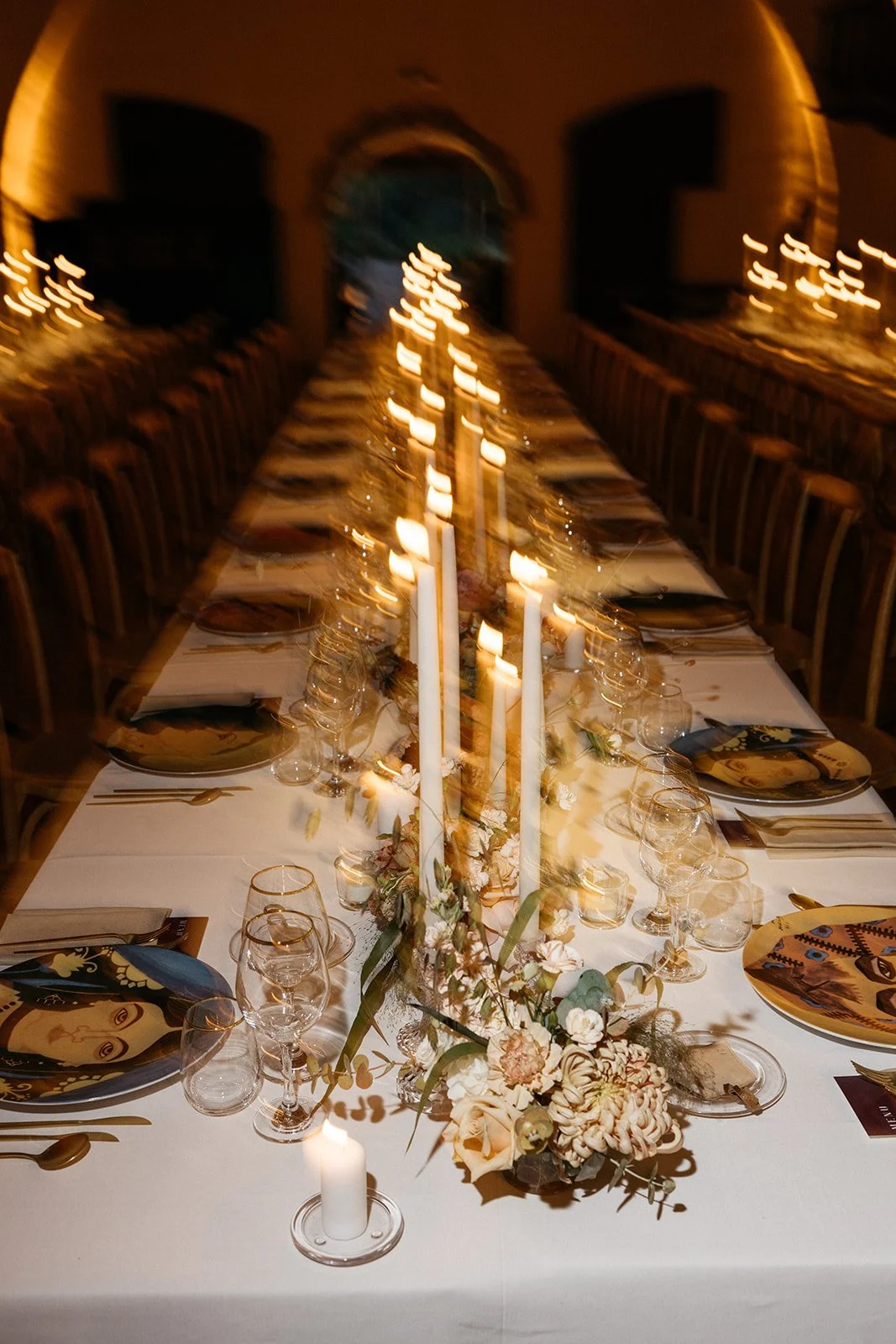 A long dining table set for a formal event, decorated with flowers, tall candles, and glassware, under warm lighting.
