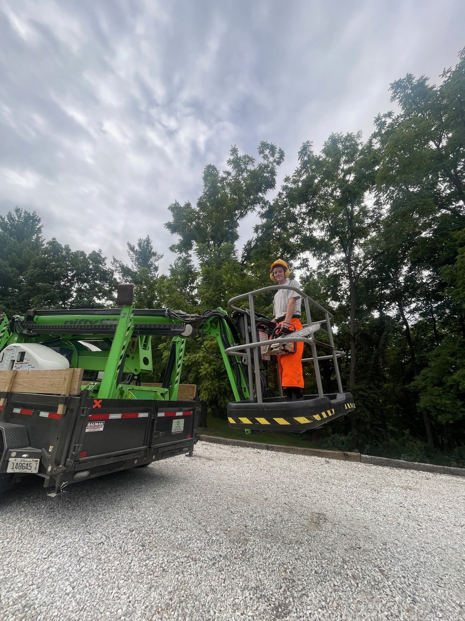 A person wearing a yellow safety helmet and orange safety pants operating a green cherry picker lift, with a textured gravel ground and trees in the background.
