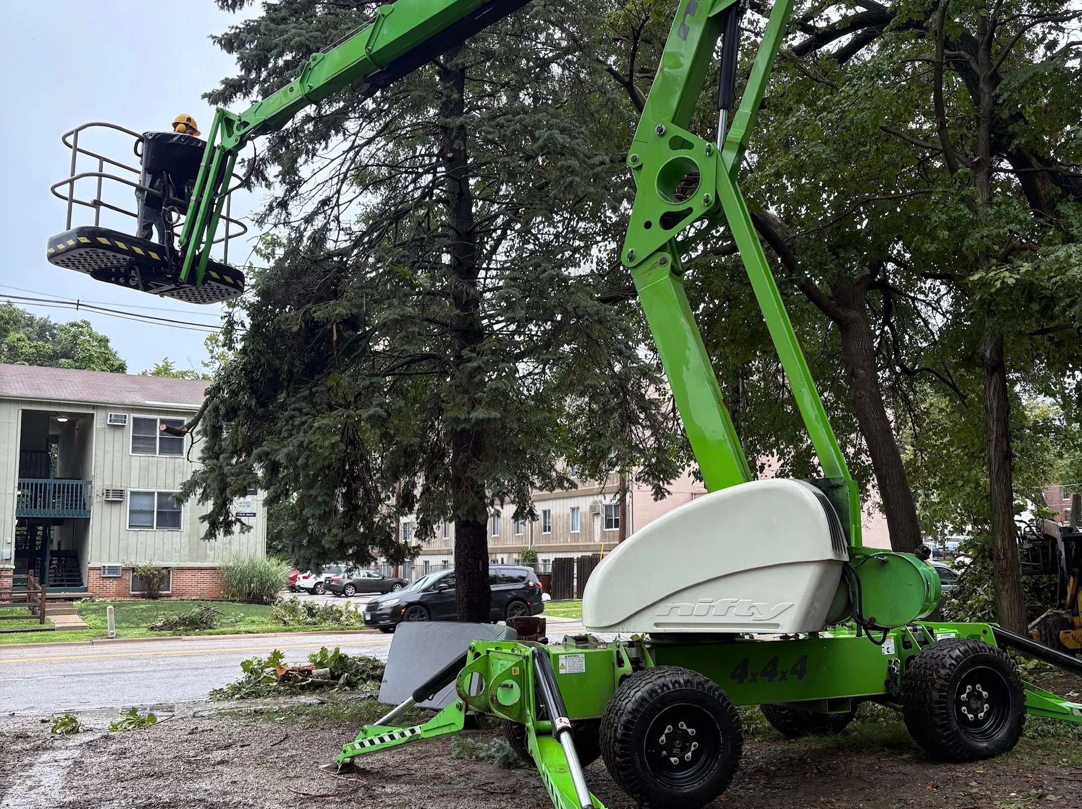 A worker operating a green cherry picker lift, trimming a tree with green foliage, in an apartment complex parking lot.
