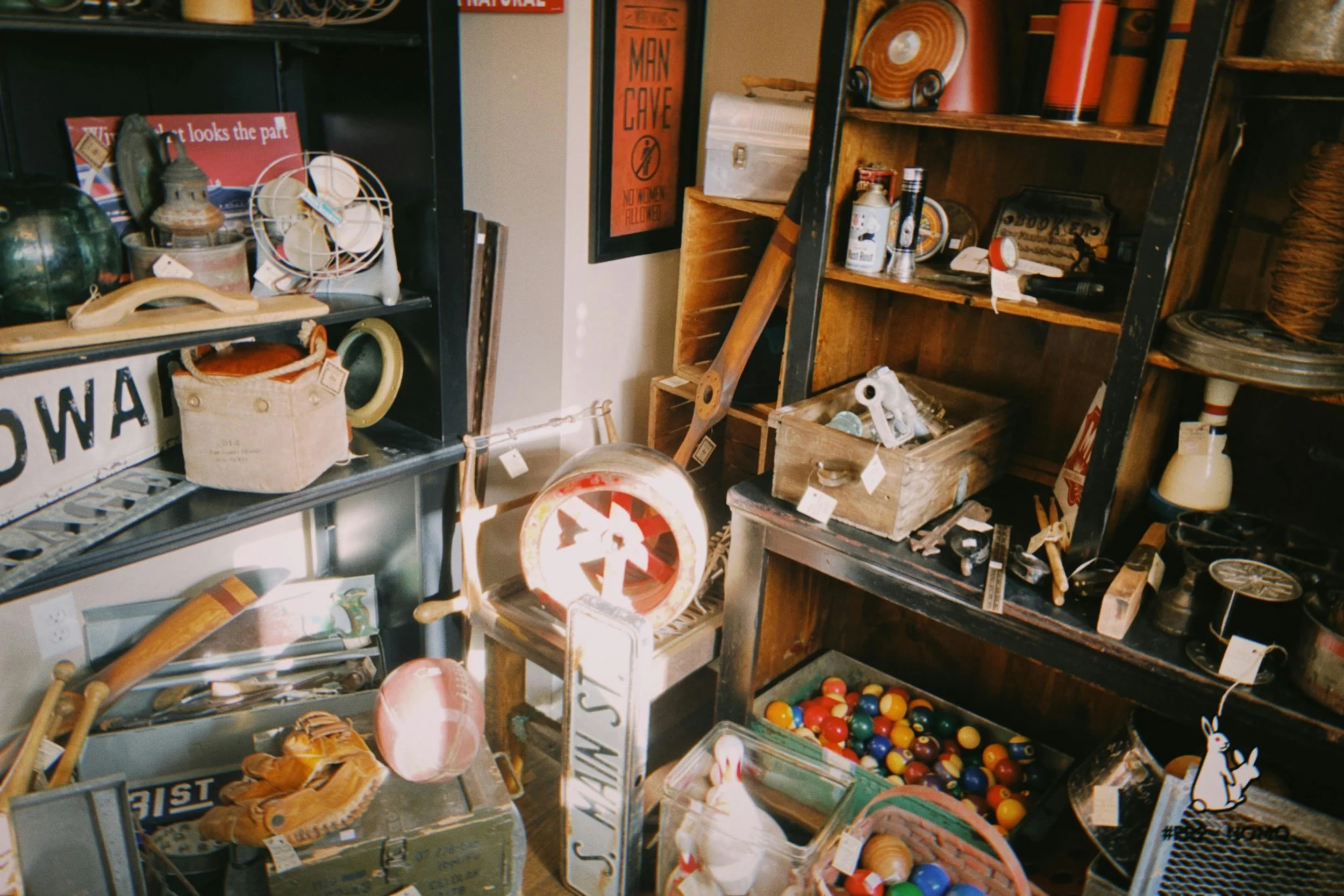 A cluttered antique shop display with various vintage items, signs, toys, and household objects arranged on shelves and tables.
