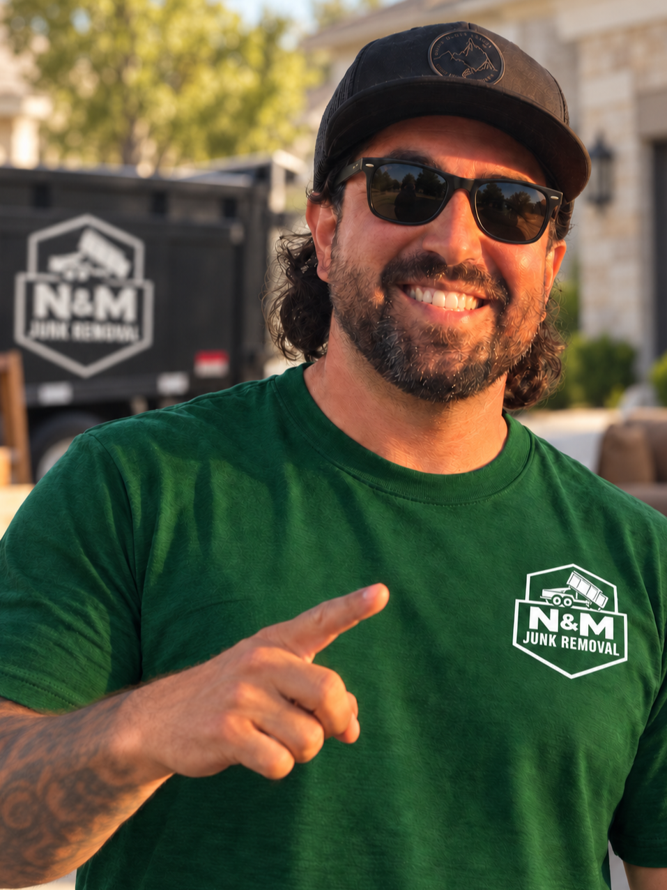 Smiling man wearing sunglasses, a green t-shirt with 'N&M Junk Removal' logo, black cap, pointing with his right index finger, outdoors with trees and a building in the background.