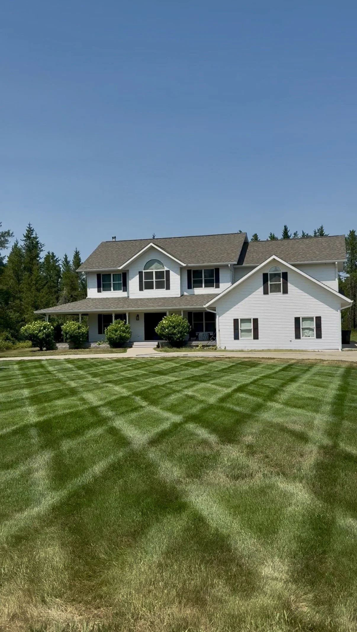 A large white two-story house with black shutters and a brown roof, surrounded by a green lawn with freshly mowed striped grass and some bushes, set against a backdrop of trees and a clear blue sky.