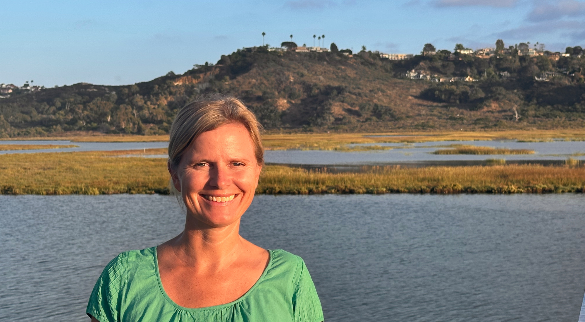 A woman with blonde hair smiling while standing near water with marshland and hills with houses in the background.