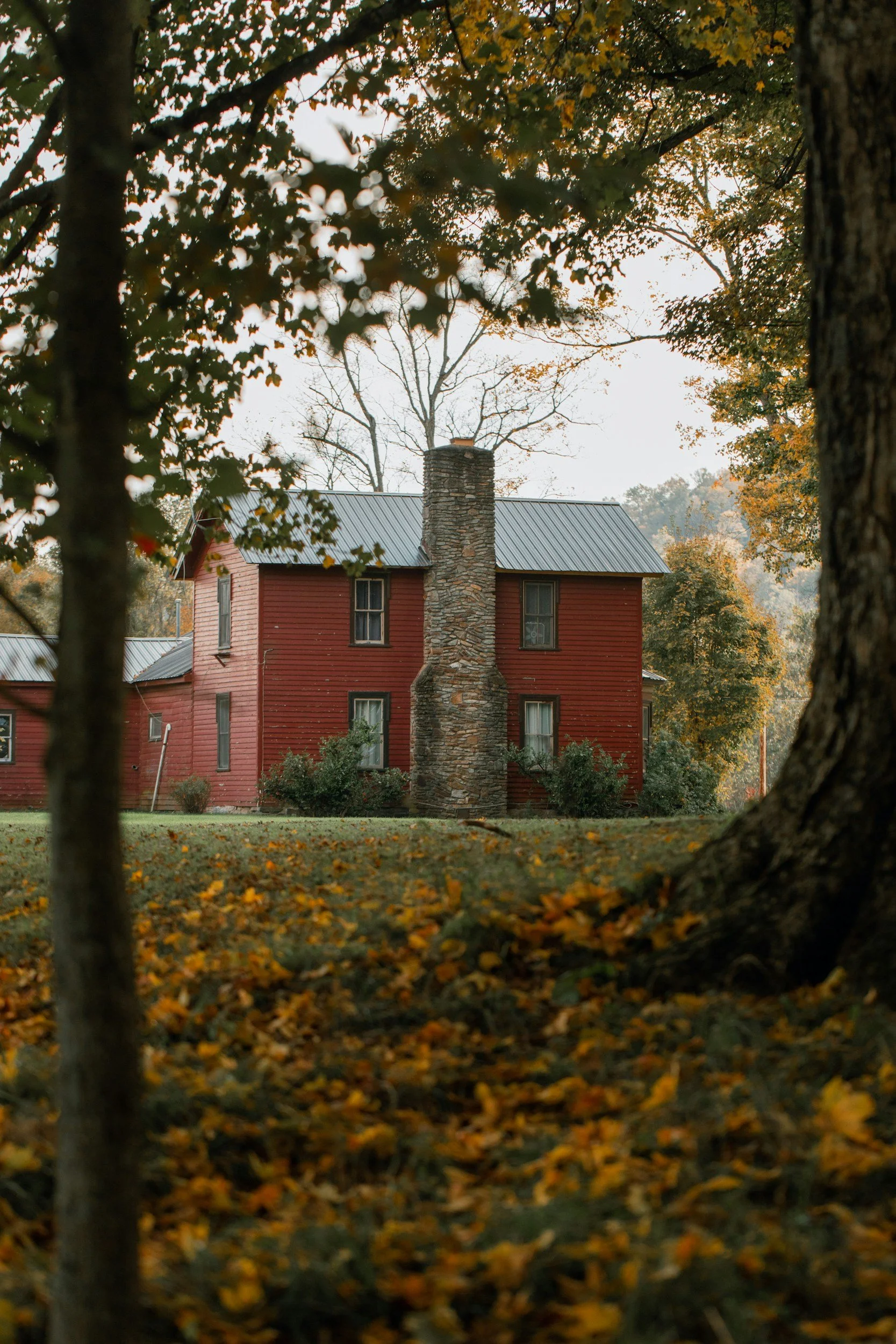 A red house with a metal roof and a tall stone chimney, surrounded by trees with yellow and orange fall leaves.