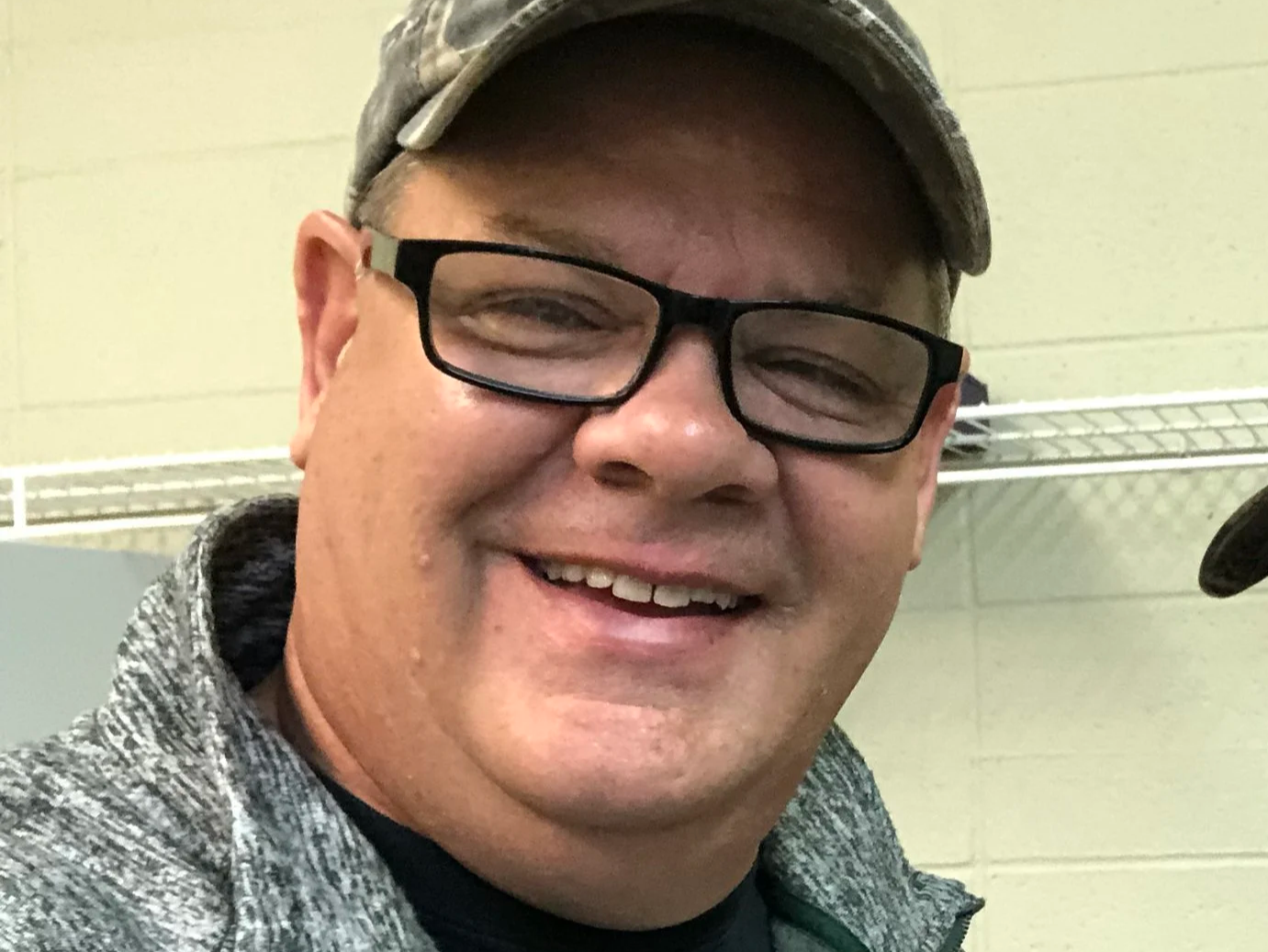 A smiling man wearing glasses and a backwards cap, standing in an indoor setting with a light green wall and wire shelving in the background.