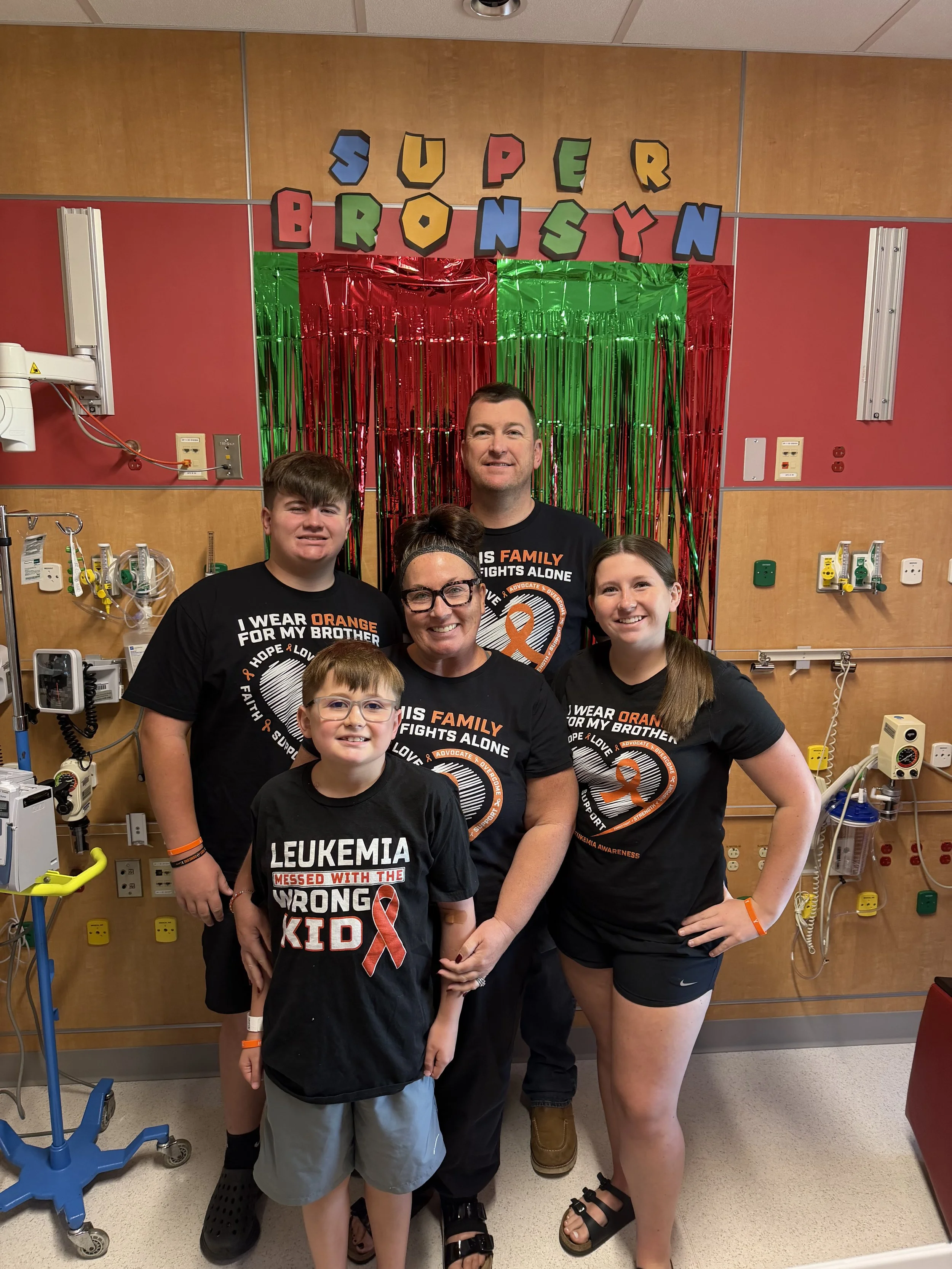 A group of six people posing in a hospital room with medical equipment, colorful decorations, and a sign that reads "Super BronySyn". The group includes two children, two women, and two men, all wearing black T-shirts related to leukemia awareness.