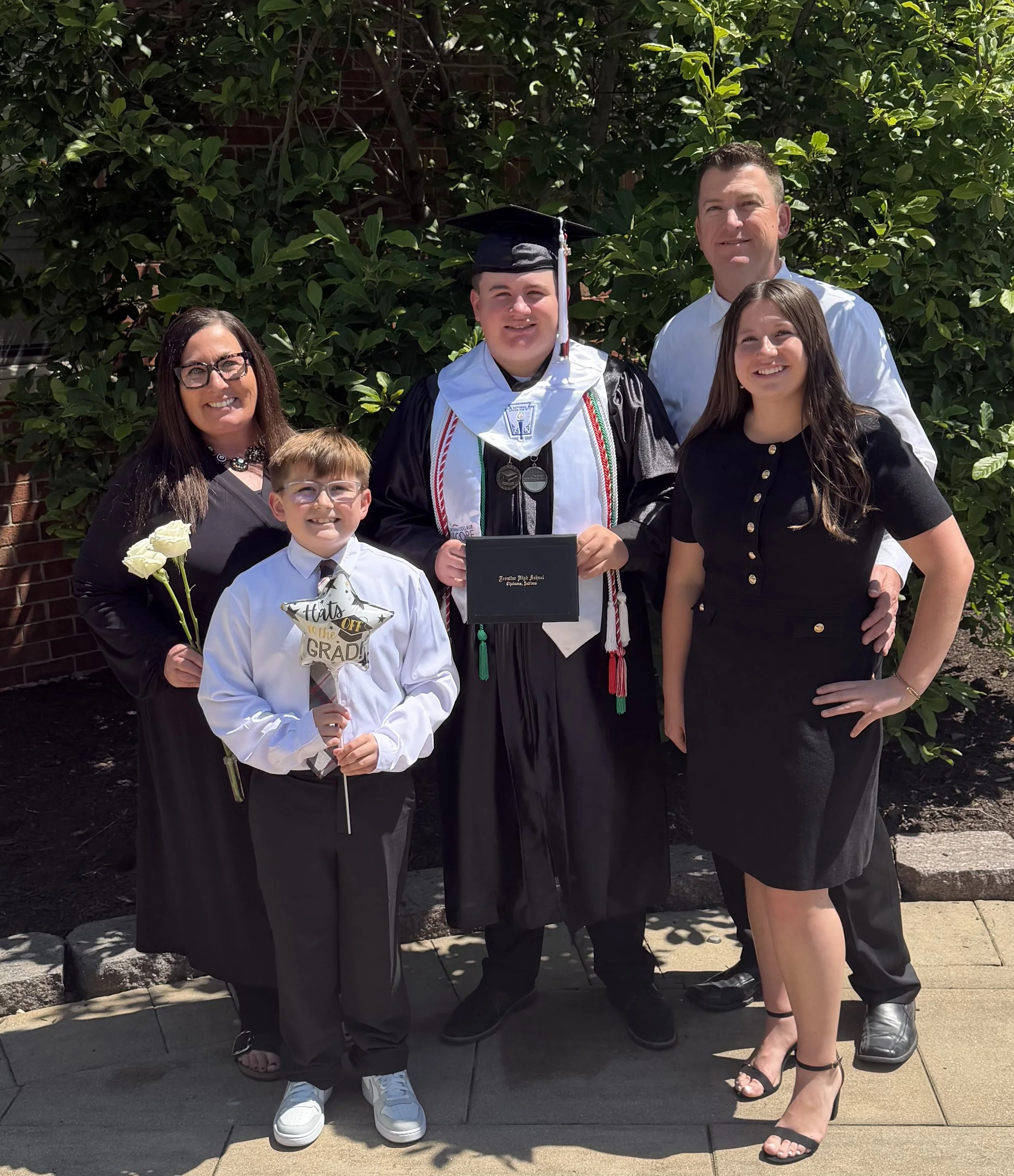 Family celebrating graduation outdoors with a young man in cap and gown holding a diploma, surrounded by family members smiling, some with flowers, against a leafy background.