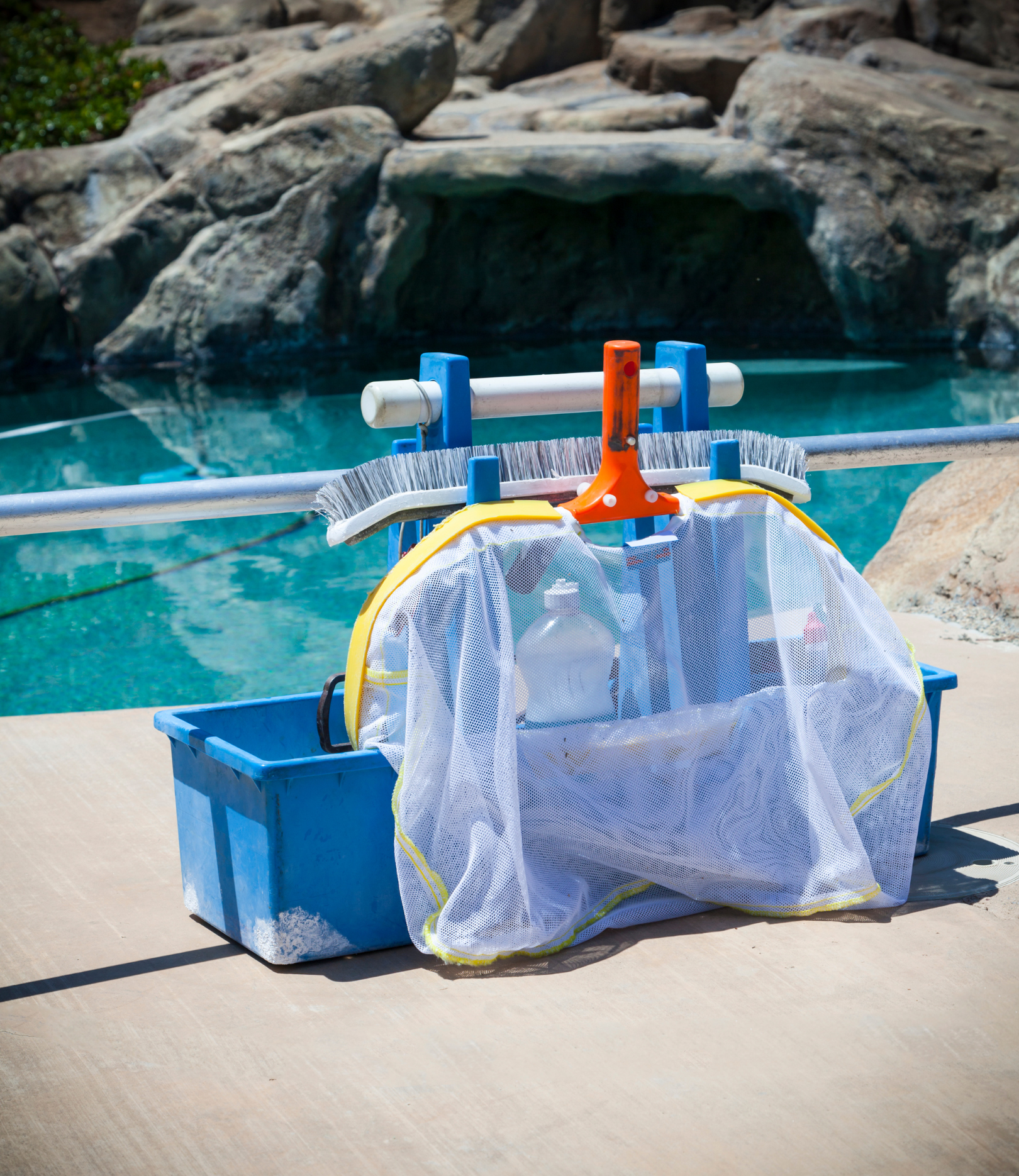 A blue plastic bin holding pool chemicals and a skimming net, placed on concrete beside a swimming pool with rocks in the background.