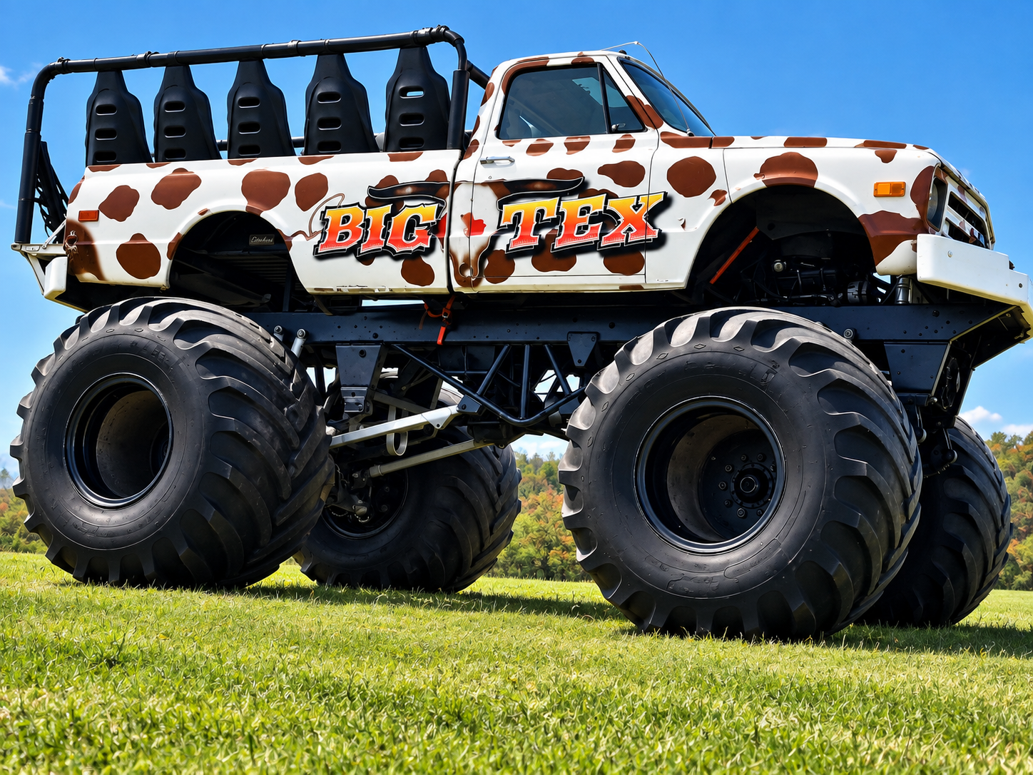 A large monster truck painted with a cow print pattern, featuring big tires and the words "Big Tex" on its side, parked on the grass under a blue sky.