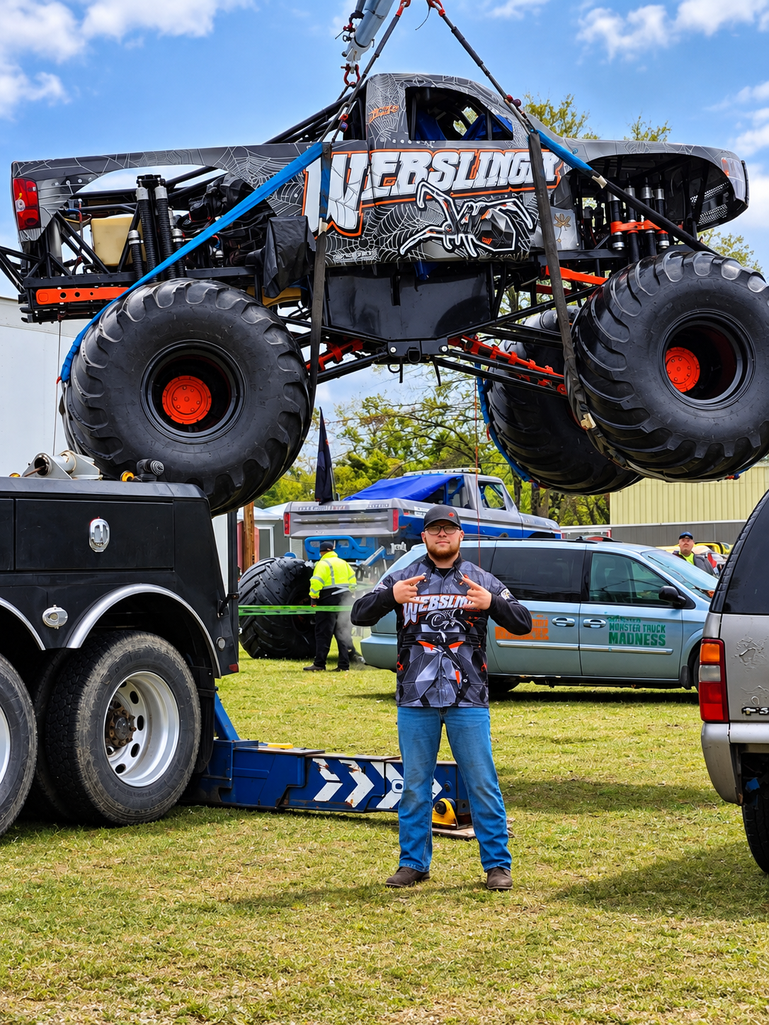A man making a gesture with his hands in front of a monster truck with large tires at an outdoor event. The truck is suspended above the ground, and there are other vehicles and people in the background.