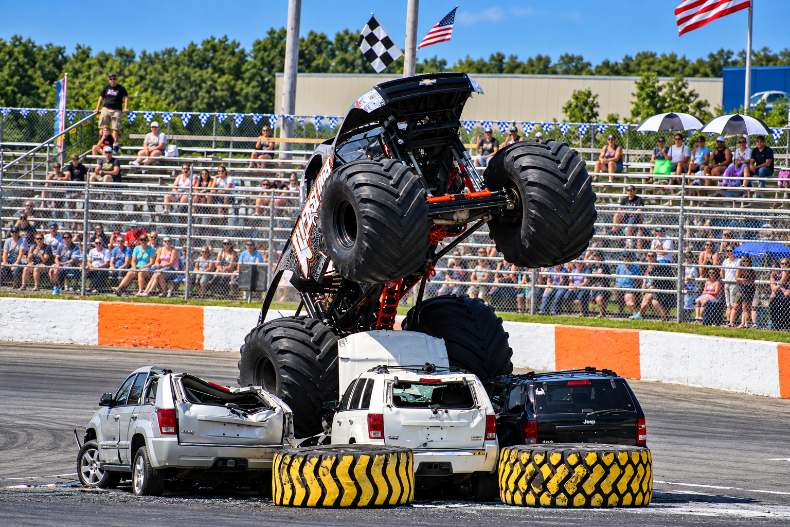 A monster truck is performing a stunt, balancing on top of three cars during a motorsport event, with spectators watching from the stands.