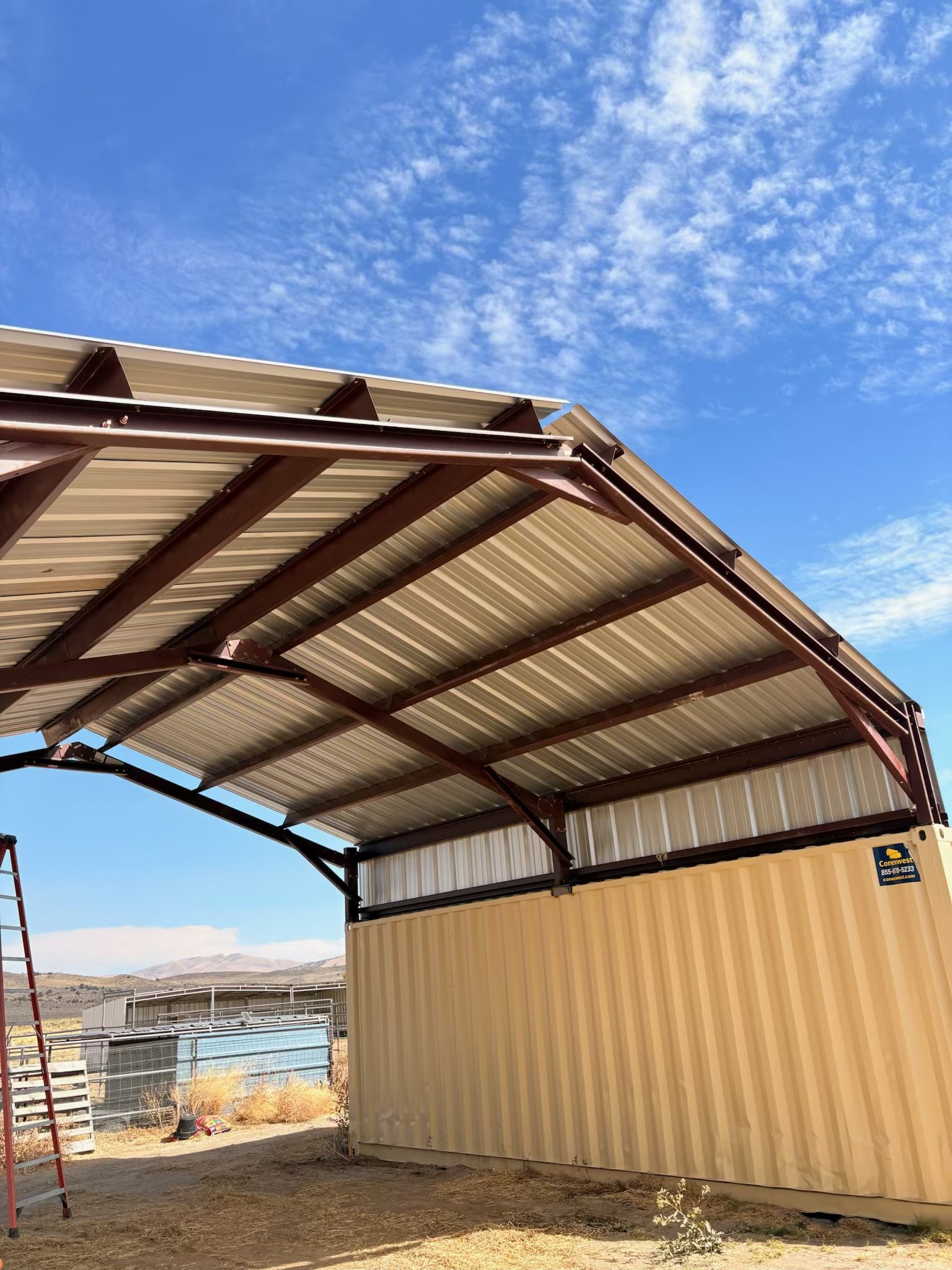 Steel-framed metal roof addition over a tan shipping container, with a ladder and desert mountains under a blue sky.