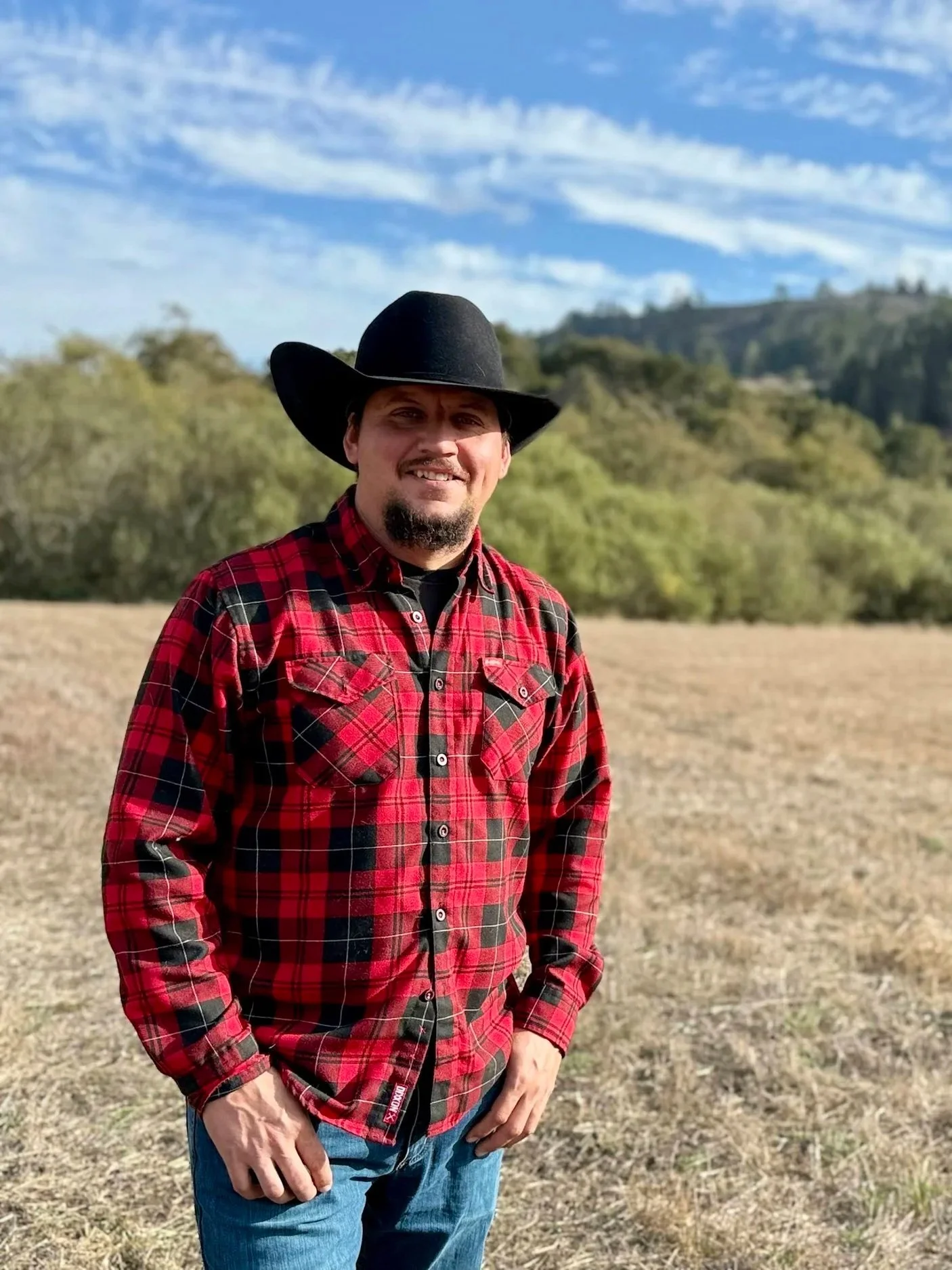 Man wearing a black cowboy hat and red plaid shirt standing in a sunny field