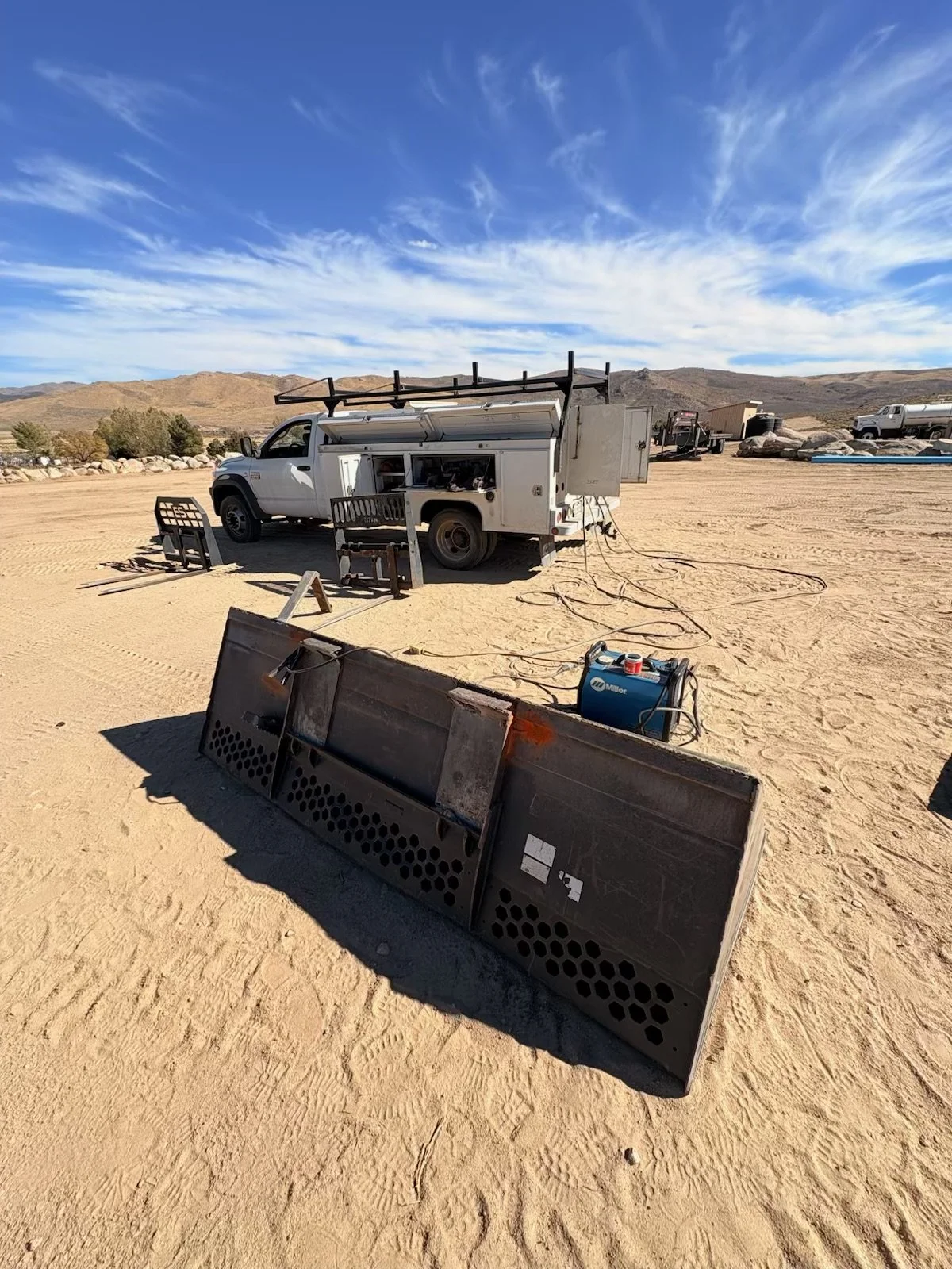 White utility truck with welding equipment and a large bulldozer blade at a sandy desert worksite