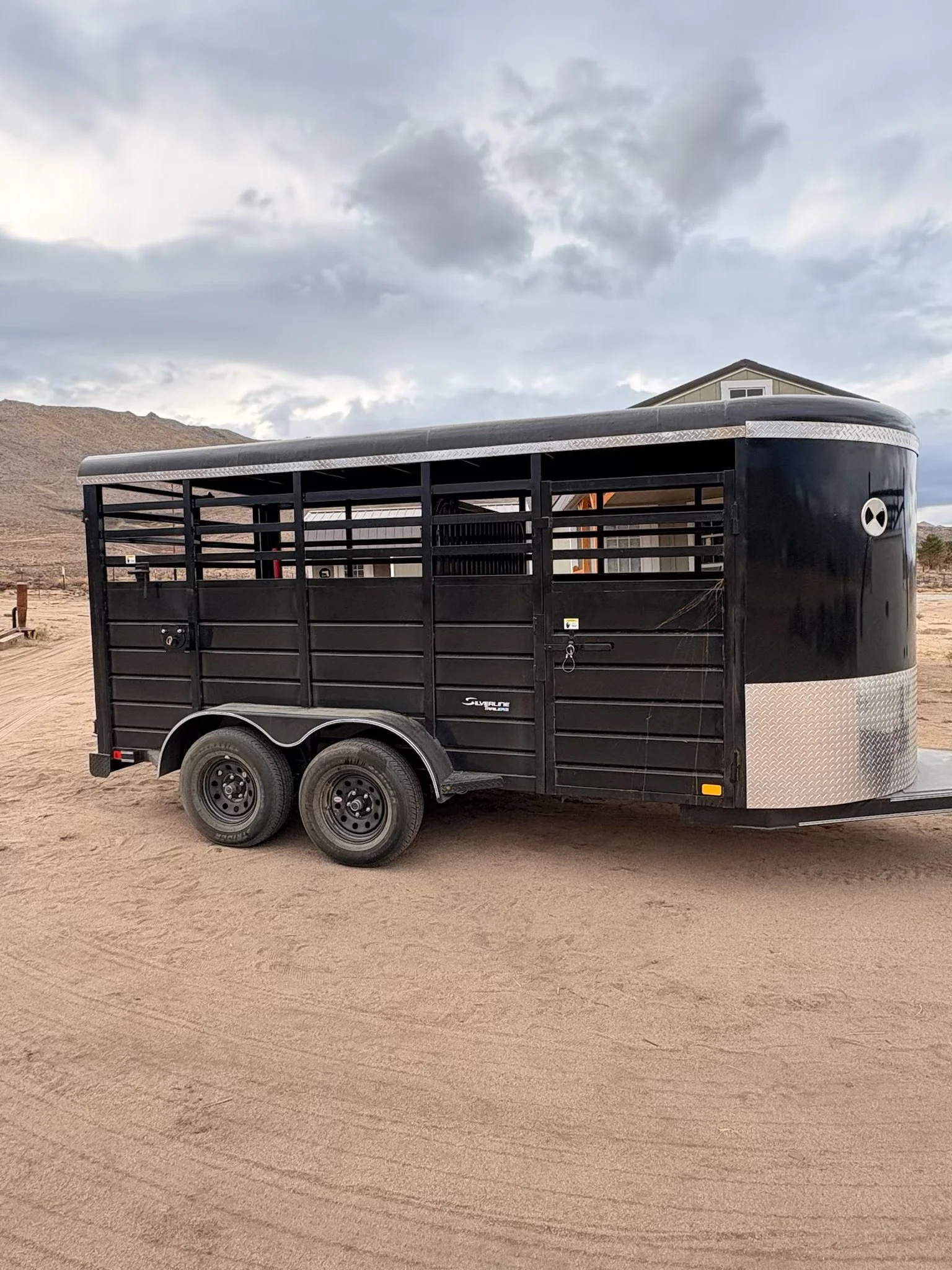 Black aluminum livestock trailer parked on sandy ground near a house and rugged mountain under cloudy skies