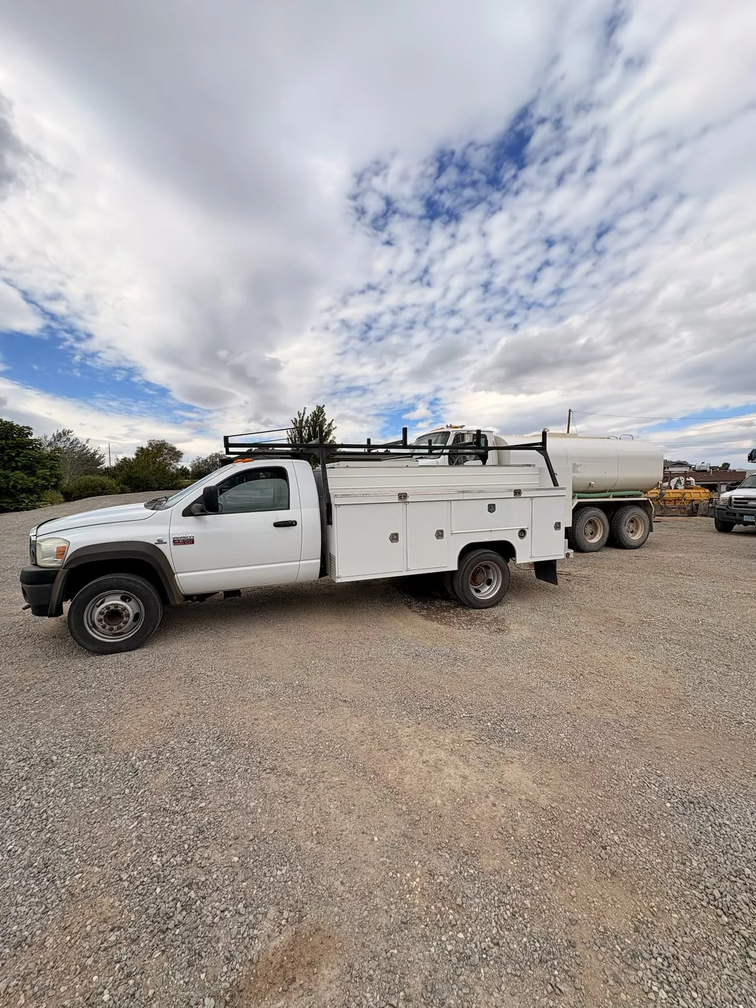 White service truck with utility bed parked on a gravel lot beside a tanker trailer under a cloudy sky