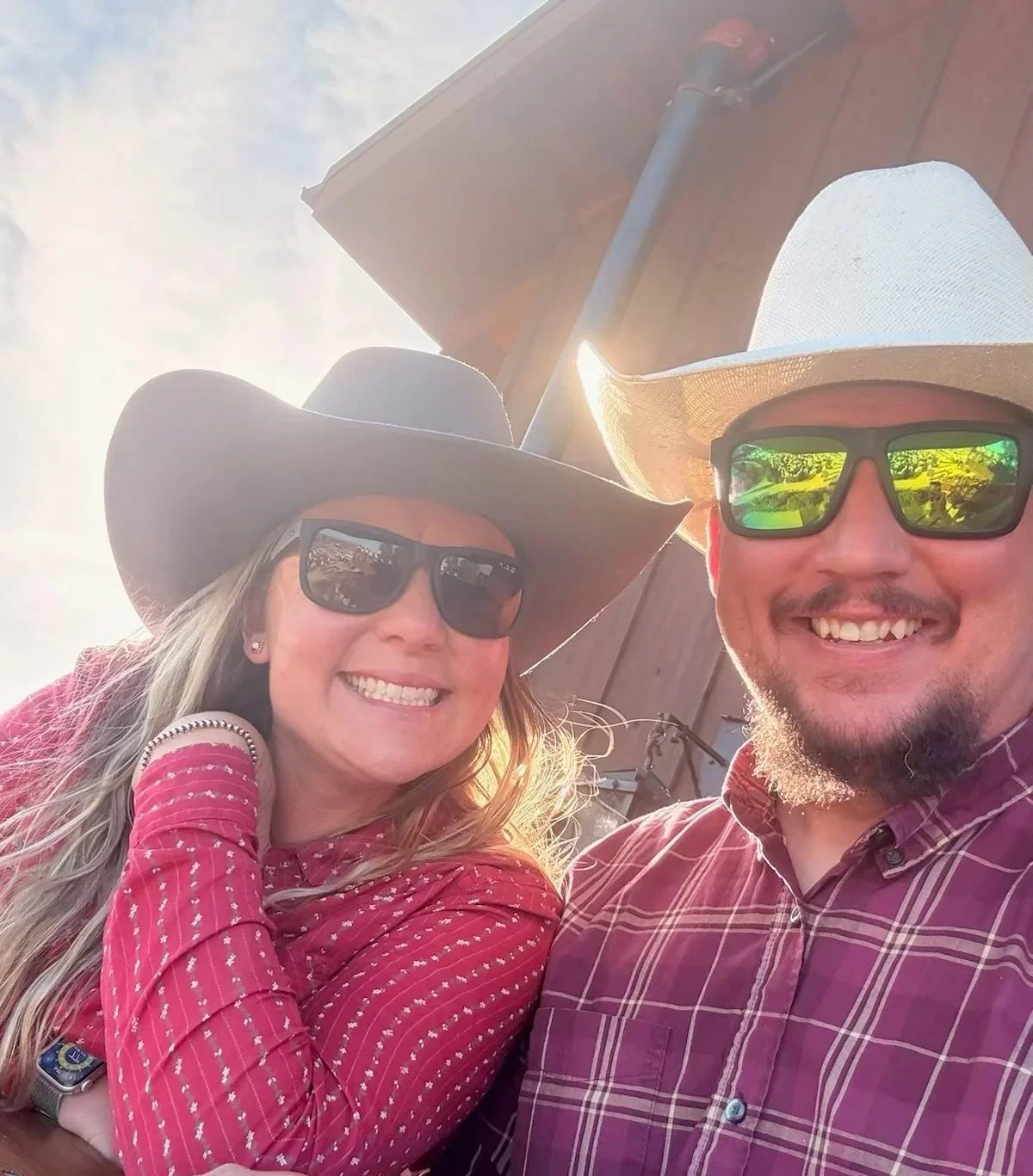 Smiling couple wearing cowboy hats and sunglasses under a patio umbrella in bright sunlight