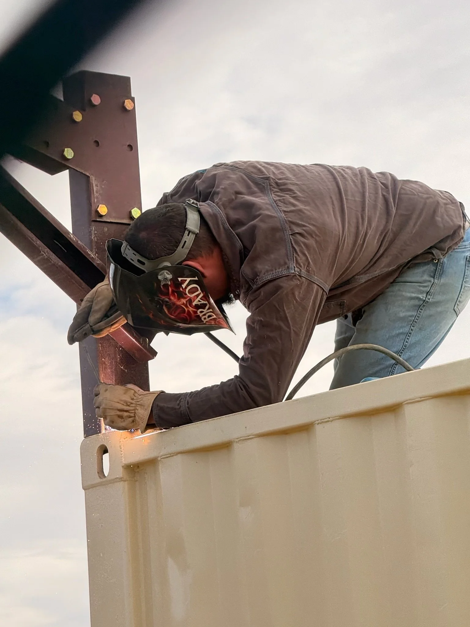Worker wearing a welding helmet and gloves repairs a metal structure from a lift platform.