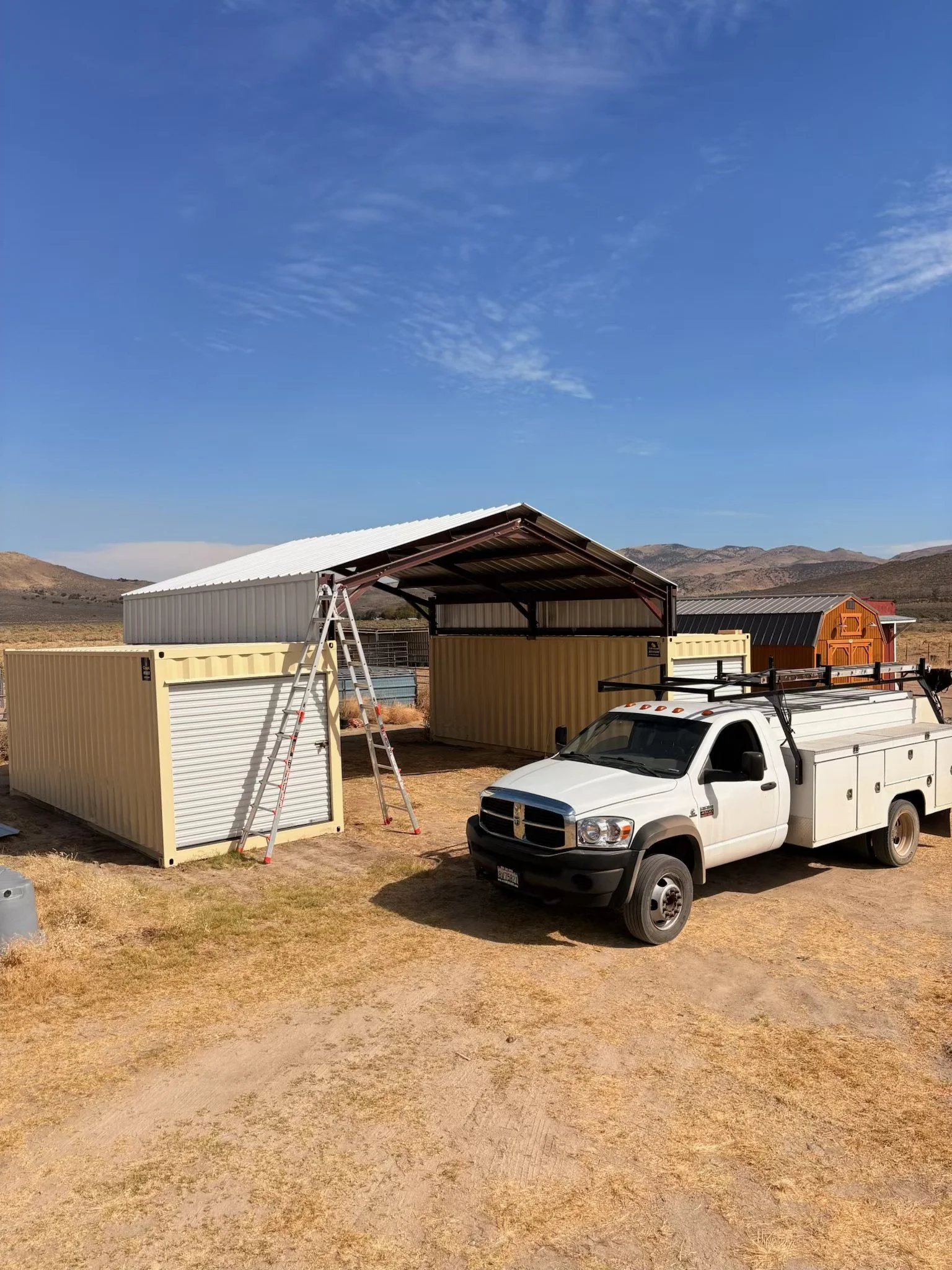 White utility truck parked beside metal storage buildings and a ladder in a dry mountainous landscape