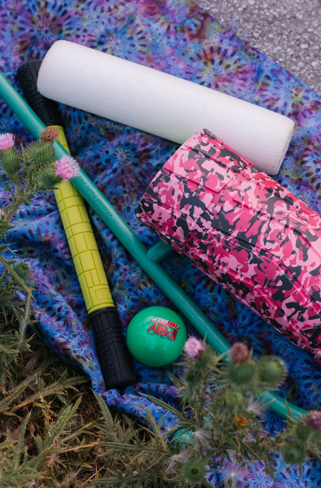 Colorful patterned blanket spread on the ground with a pink camo-patterned box, a foam roller, a blue and green pole, a green ball, and some small pink flowers.