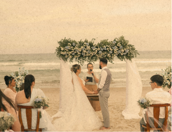 Beach wedding ceremony with a bride and groom standing under a floral arch, facing each other, during their wedding vows, with guests seated on wooden chairs nearby.