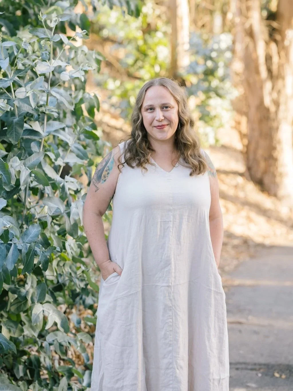 A woman with wavy light brown hair smiling, wearing a sleeveless white dress, standing outdoors on a sunny day next to green leafy bushes and trees.