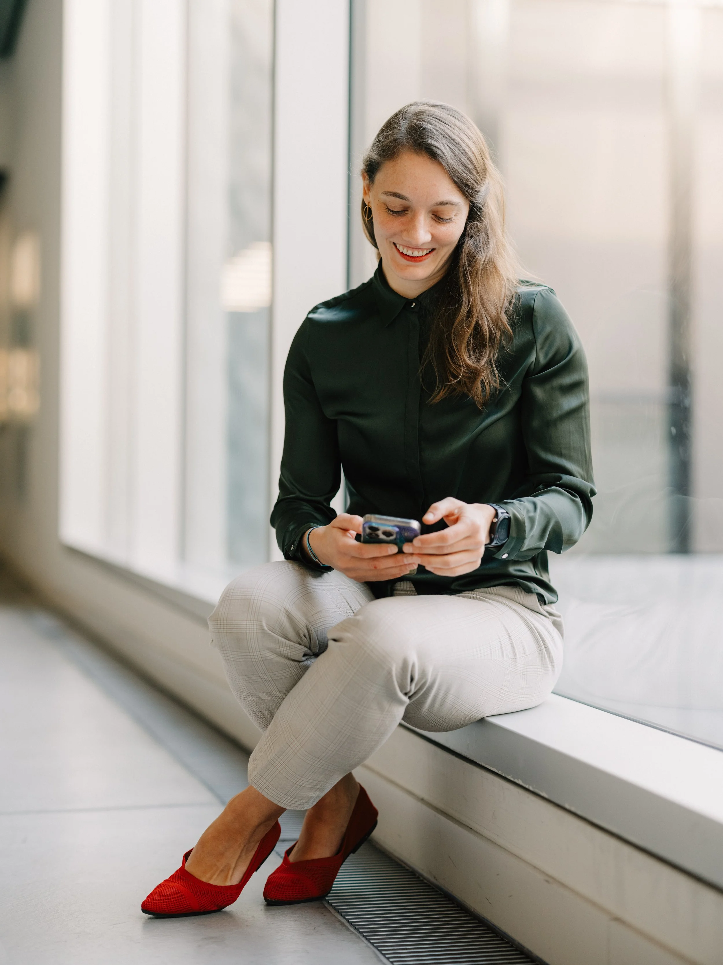 A woman wearing a green blouse, beige plaid trousers, and red shoes, sitting by a window, smiling and looking at her phone.