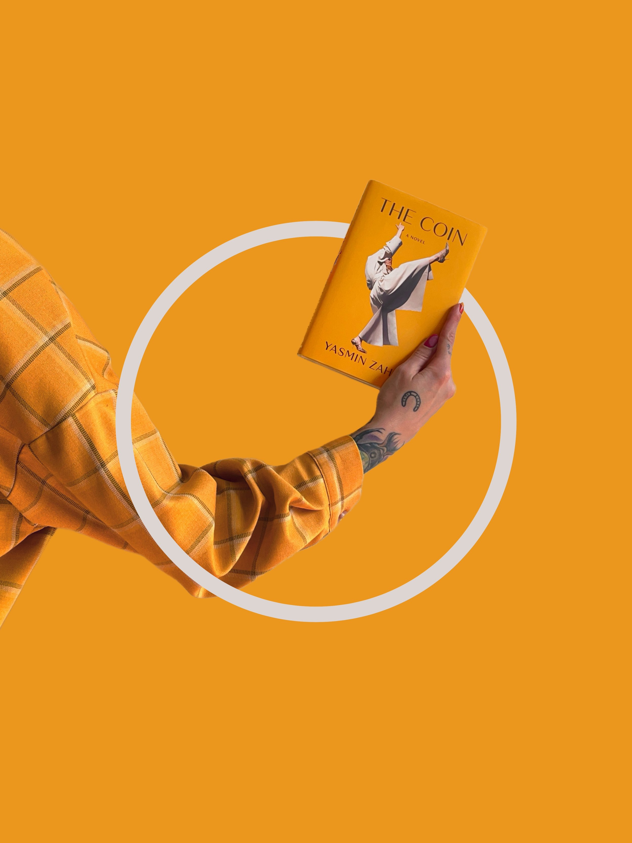 A person with tattoos on their hand and wrist holding a yellow book titled 'The Coin' by Yasmin Zahra, with a woman in white traditional attire on the cover, against an orange background with a white circle.