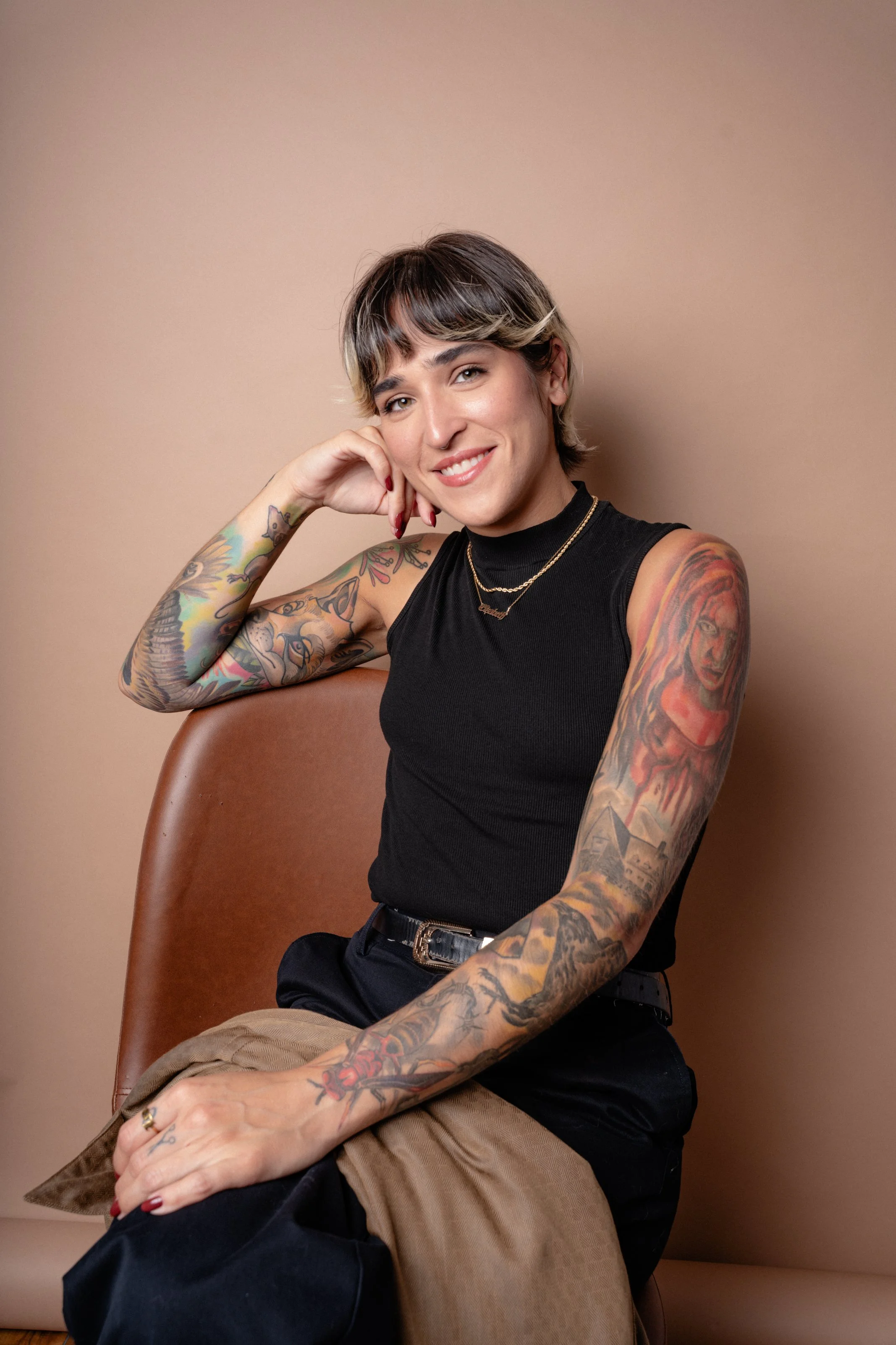A woman with short hair and tattoos on her arms is sitting on a brown chair, smiling, and resting her head on her hand against a beige background.