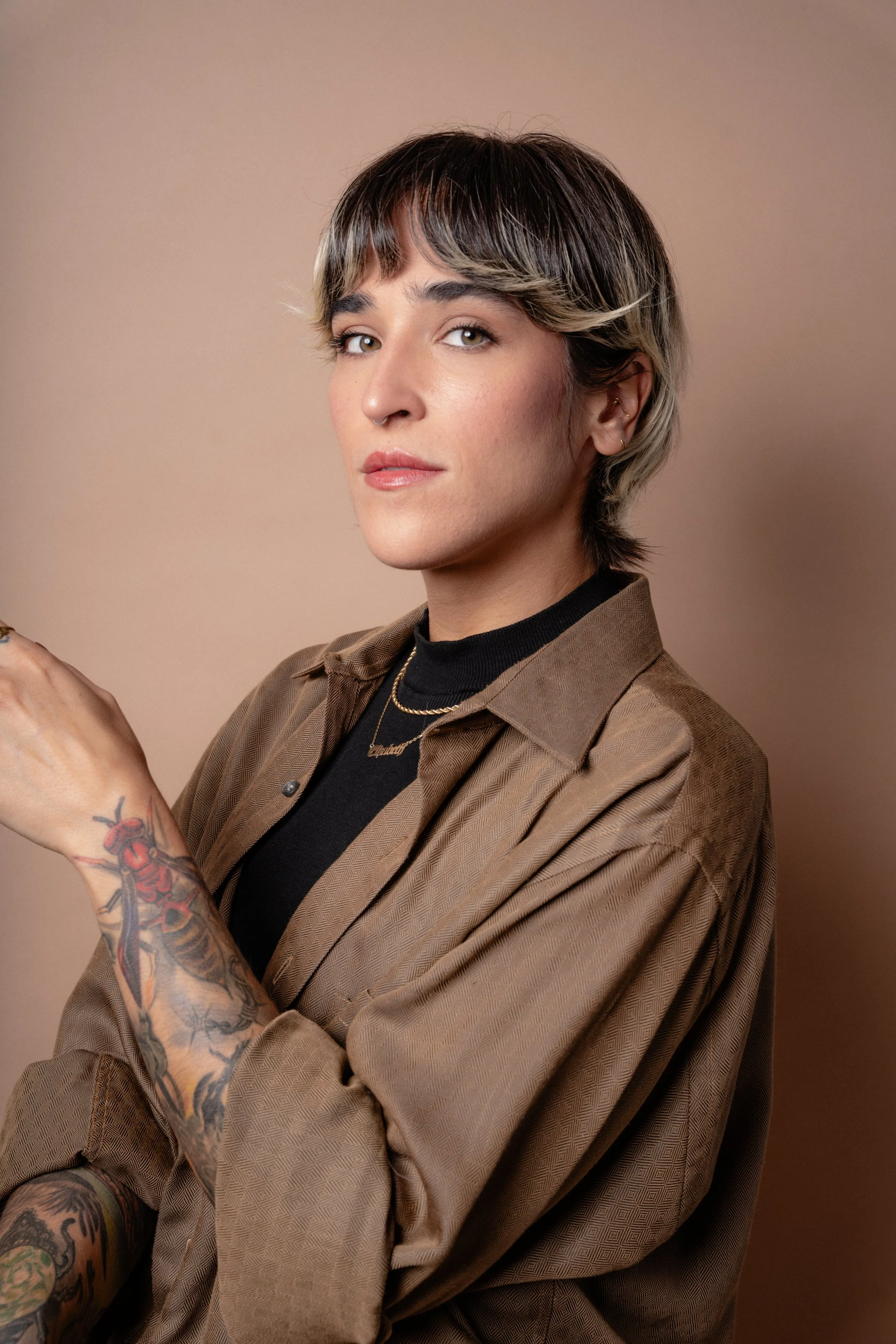 Close-up of a woman with short, layered hair, wearing a brown blazer over a black turtleneck, with tattoos on her arm, earrings, and a necklace, against a beige background.