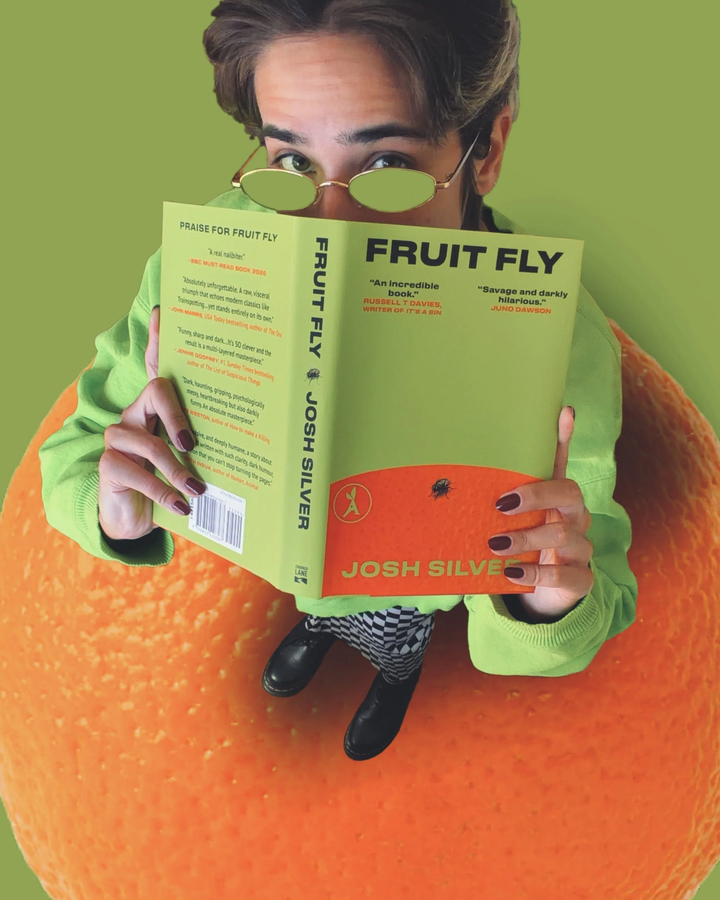 A person holding an orange book titled 'Fruit Fly' with sunglasses on, sitting on an orange textured surface.