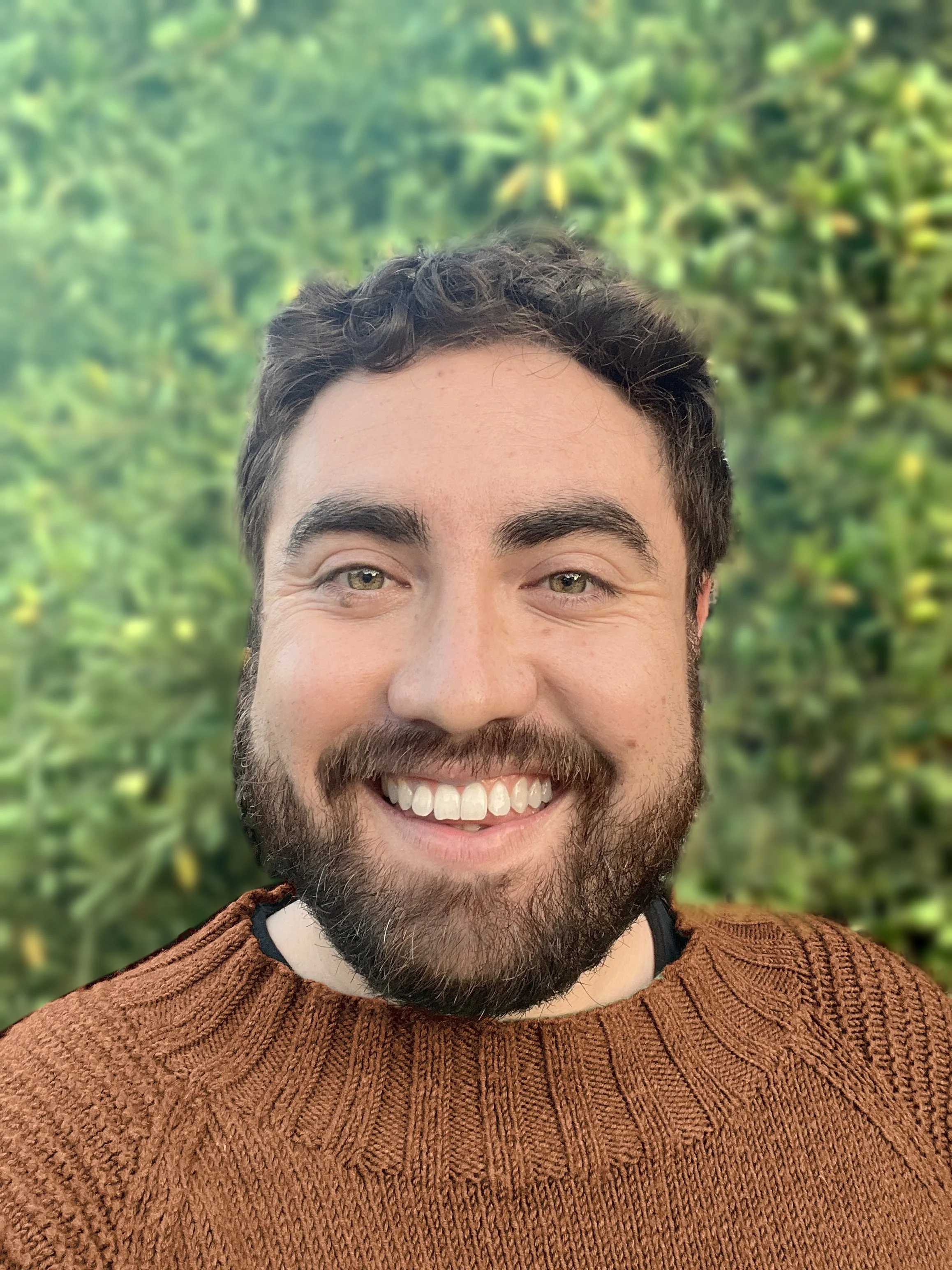 Psychodynamic and ACT psychotherapist Brewster Parsons with dark hair and a beard, wearing a rust-colored knitted sweater, standing outdoors with green foliage in the background.