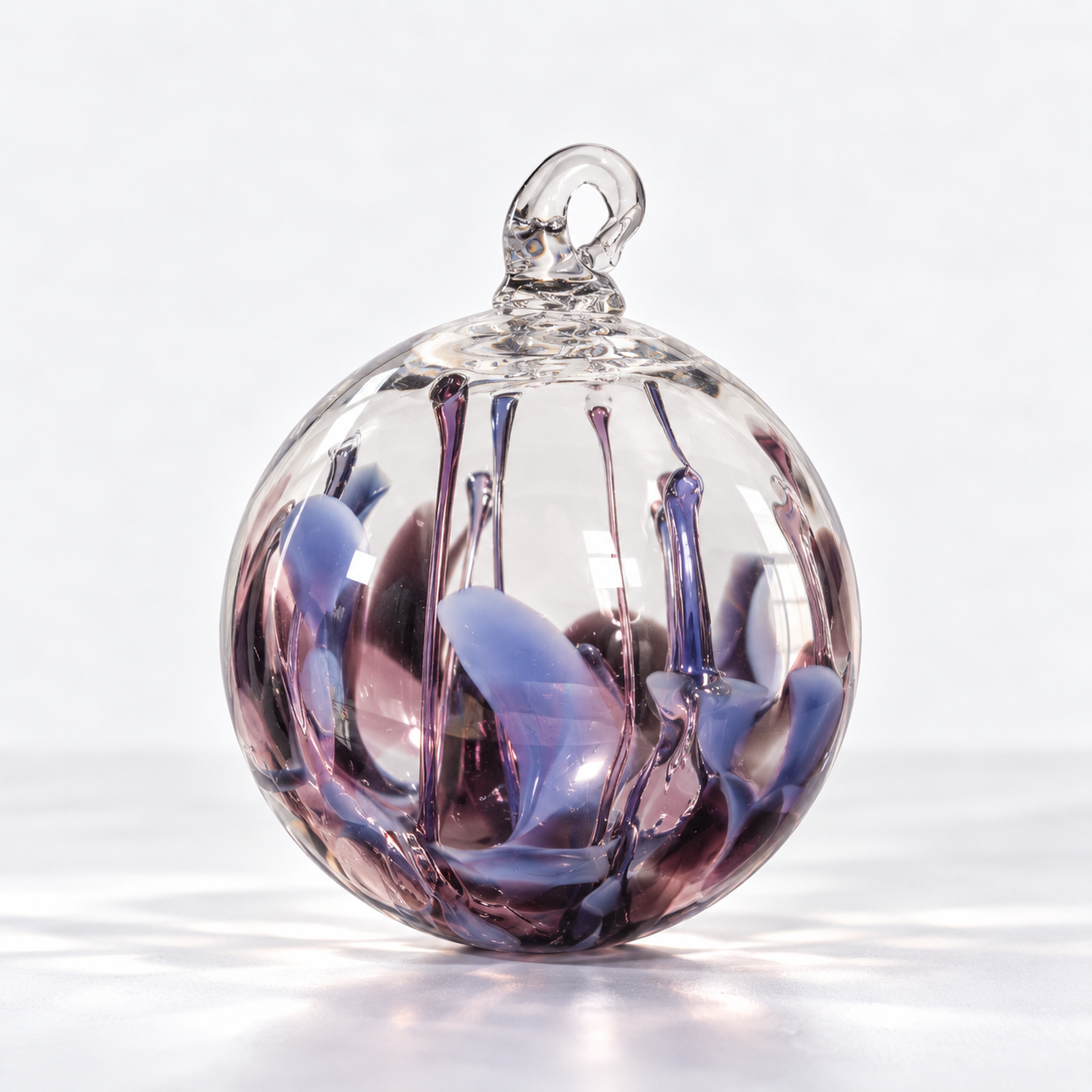 A clear glass ornament with purple and blue swirling patterns inside, topped with a small loop for hanging, against a plain white background.
