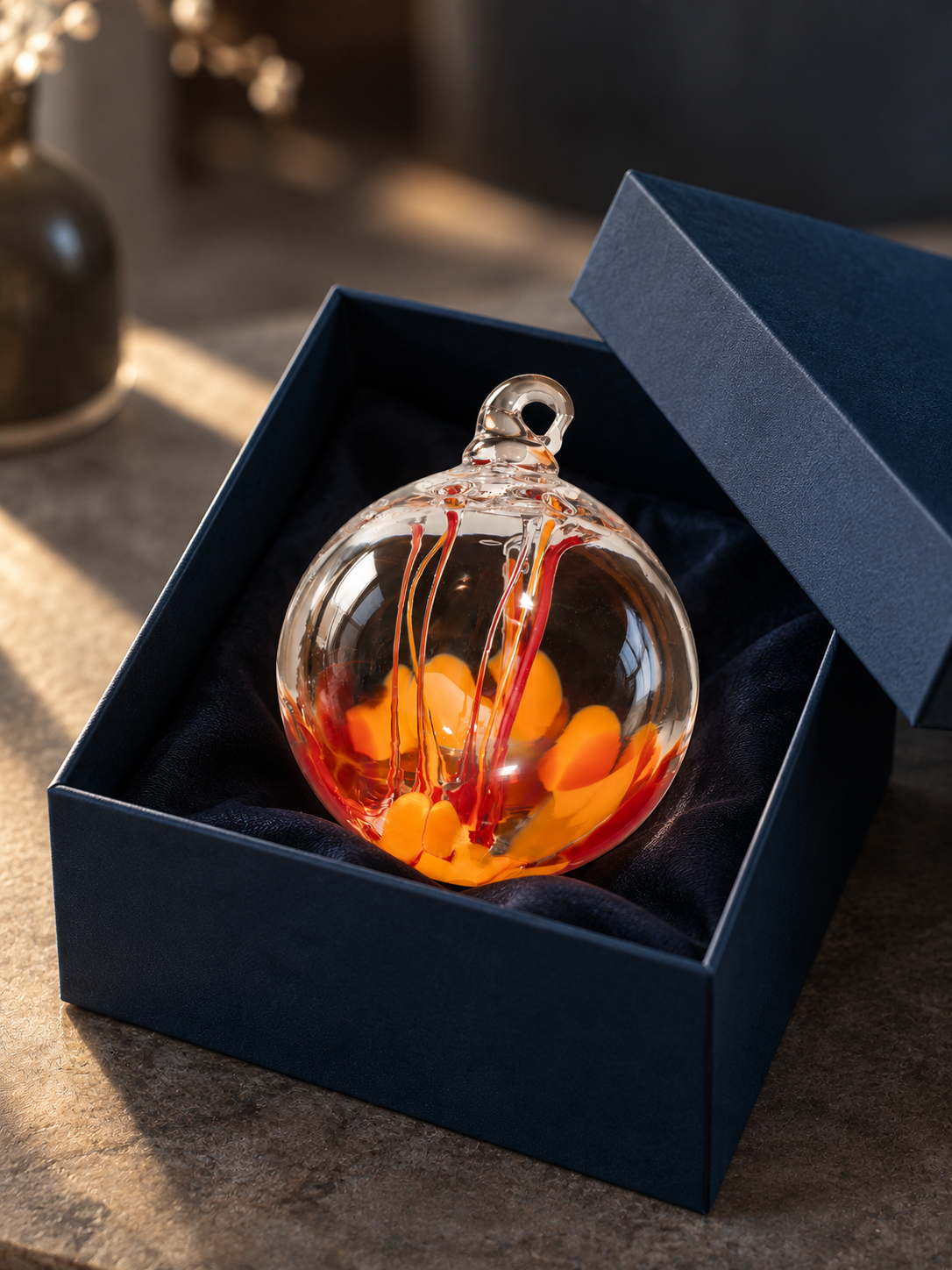 Decorative glass ornament with orange, red, and yellow swirls inside a navy blue box lined with black fabric, placed on a wooden surface.