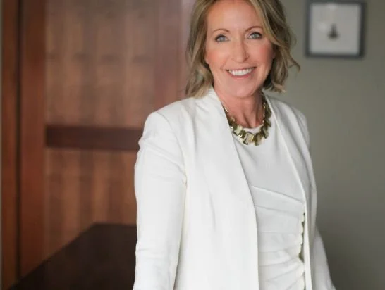 A smiling woman with blonde hair, wearing a white blazer and a statement necklace, standing indoors with a wooden wall and framed picture in the background.