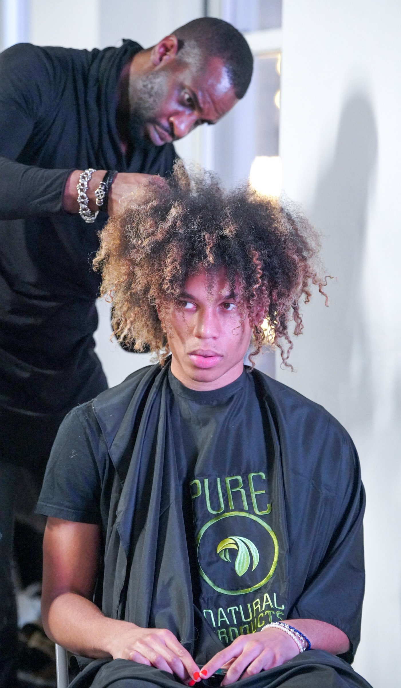 A person with curly hair sitting in a chair, wearing a black shirt with green text and a black cape, with a stylist standing behind them, styling their hair. Backstage at Laurence Basse runway show.