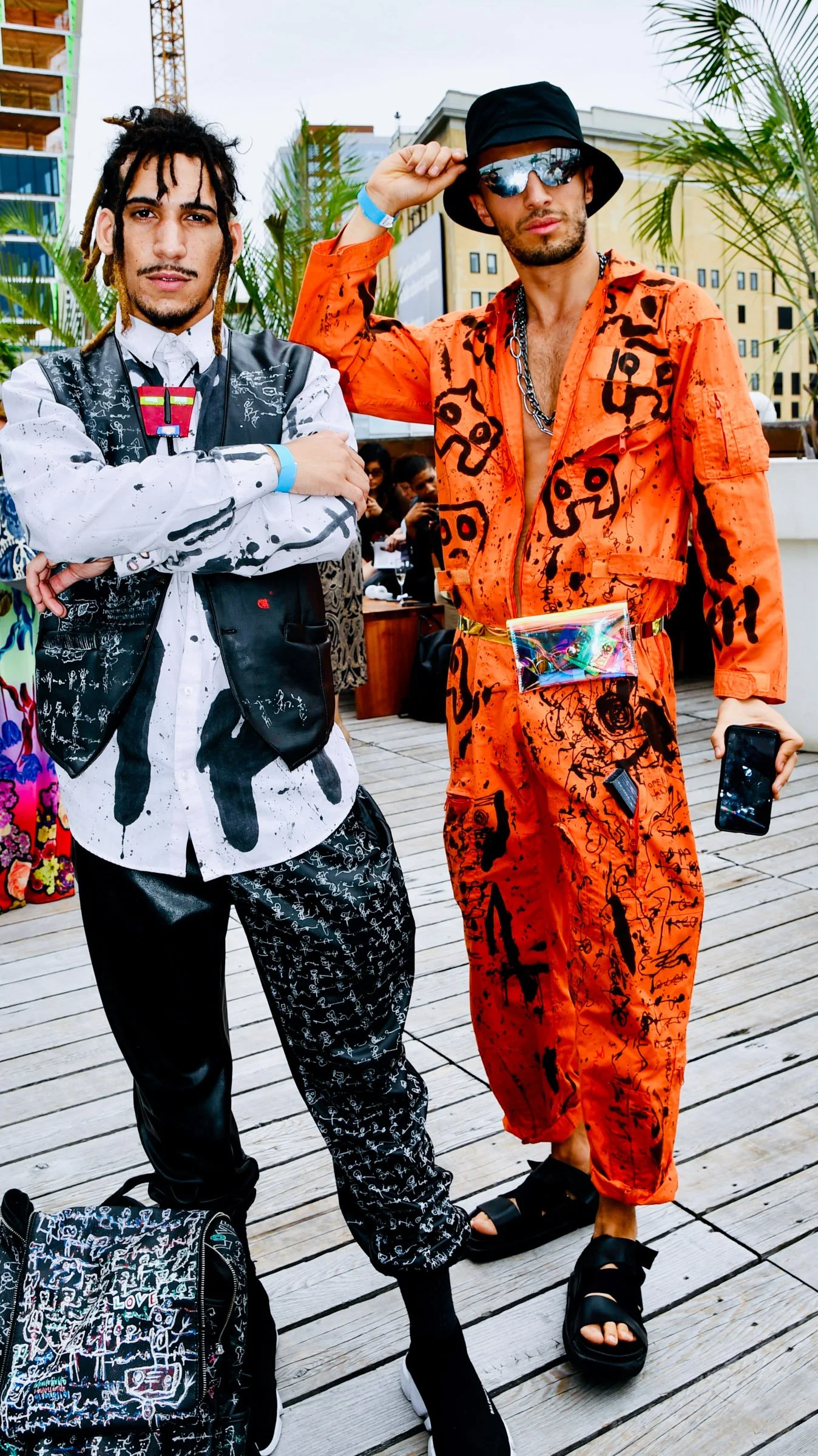 Two men with distinctive fashion styles posing outdoors. The man on the left wears a white shirt with black paint splatters, a black vest with white markings, and matching pants, with a backpack featuring graffiti-like designs. The man on the right w