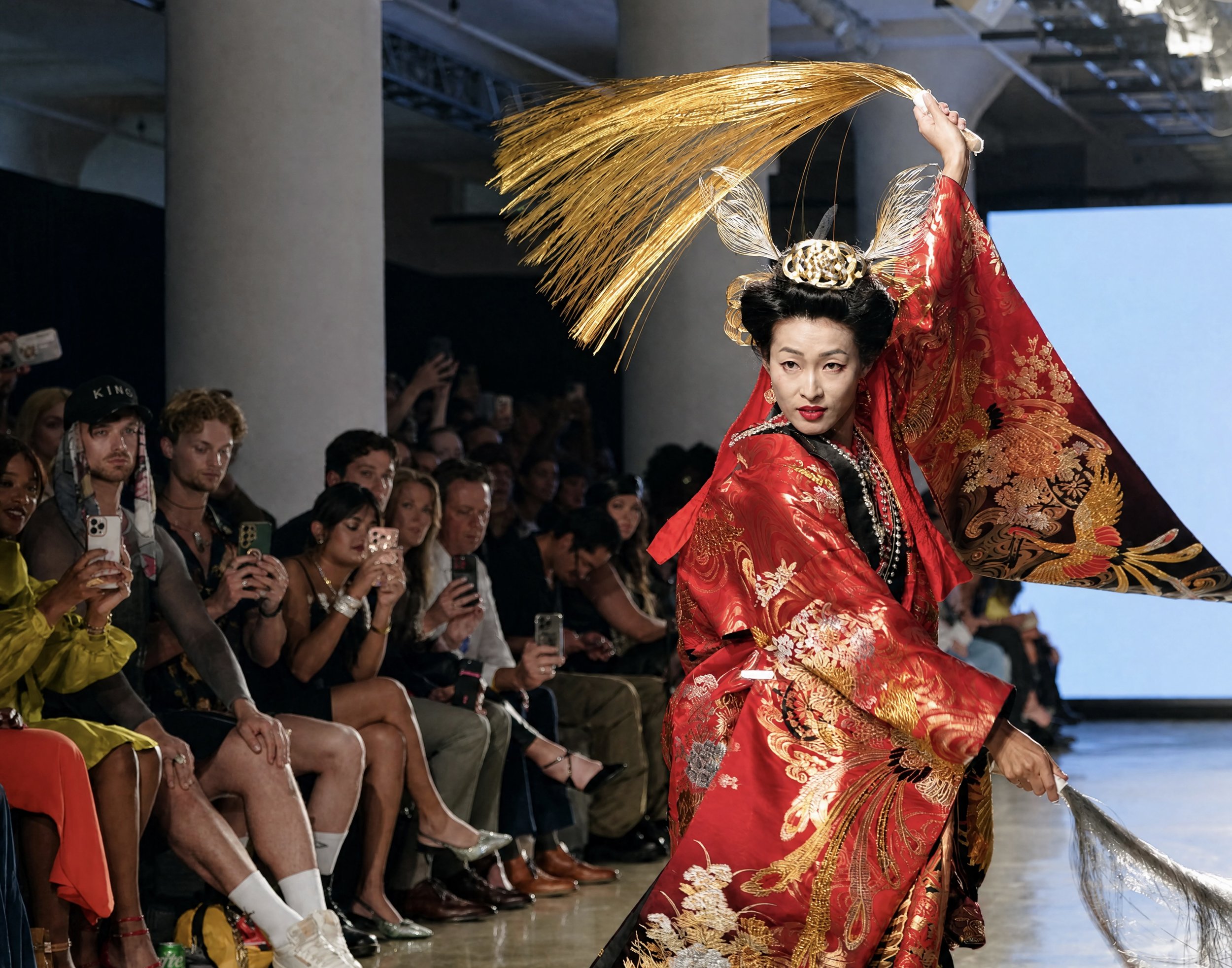 A model in a red and gold traditional Asian costume performing on a runway during a fashion show, with an audience watching and taking photos.