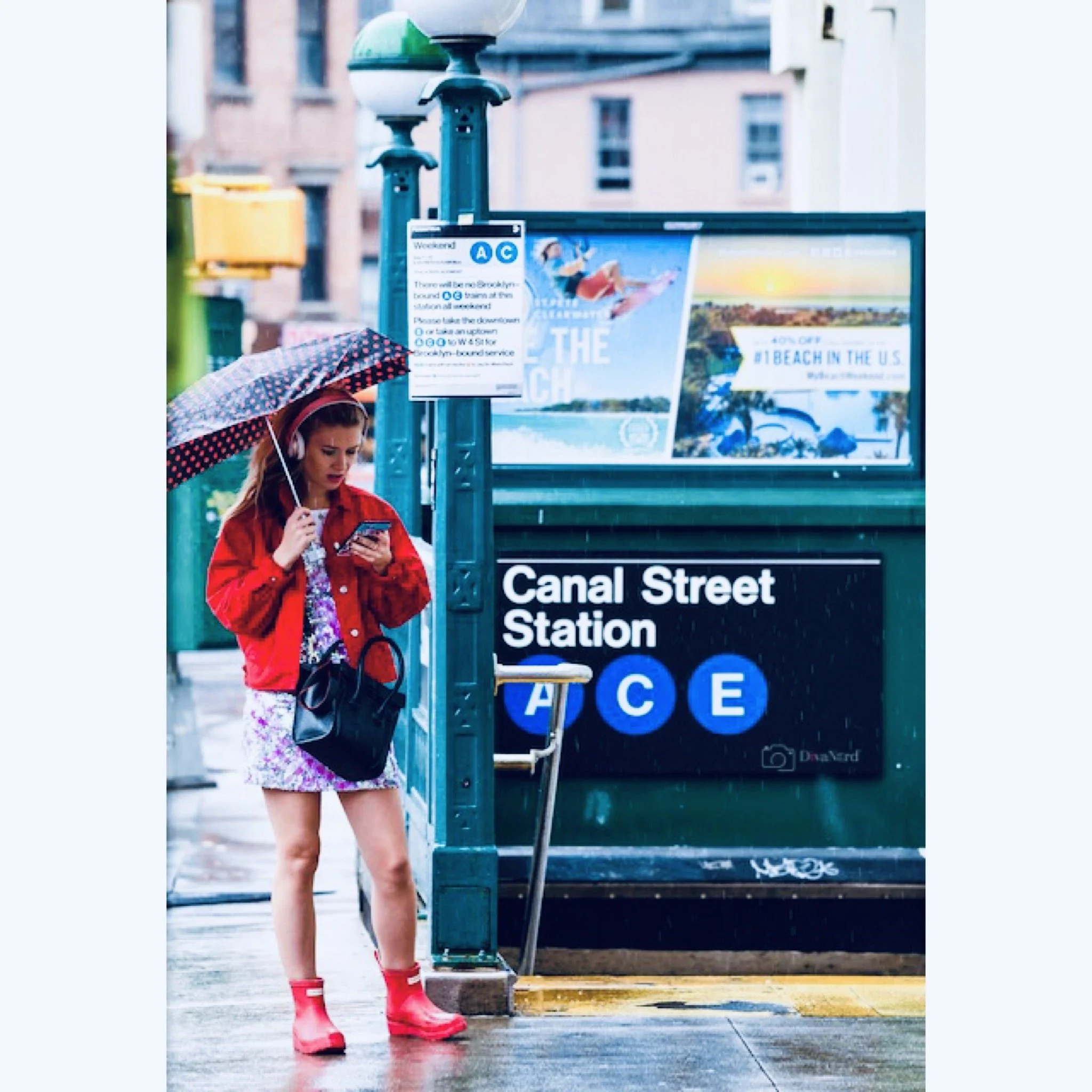 A young woman in a red jacket and floral dress, wearing red rain boots, standing at a bus stop on a rainy day, looking at her phone under an umbrella.