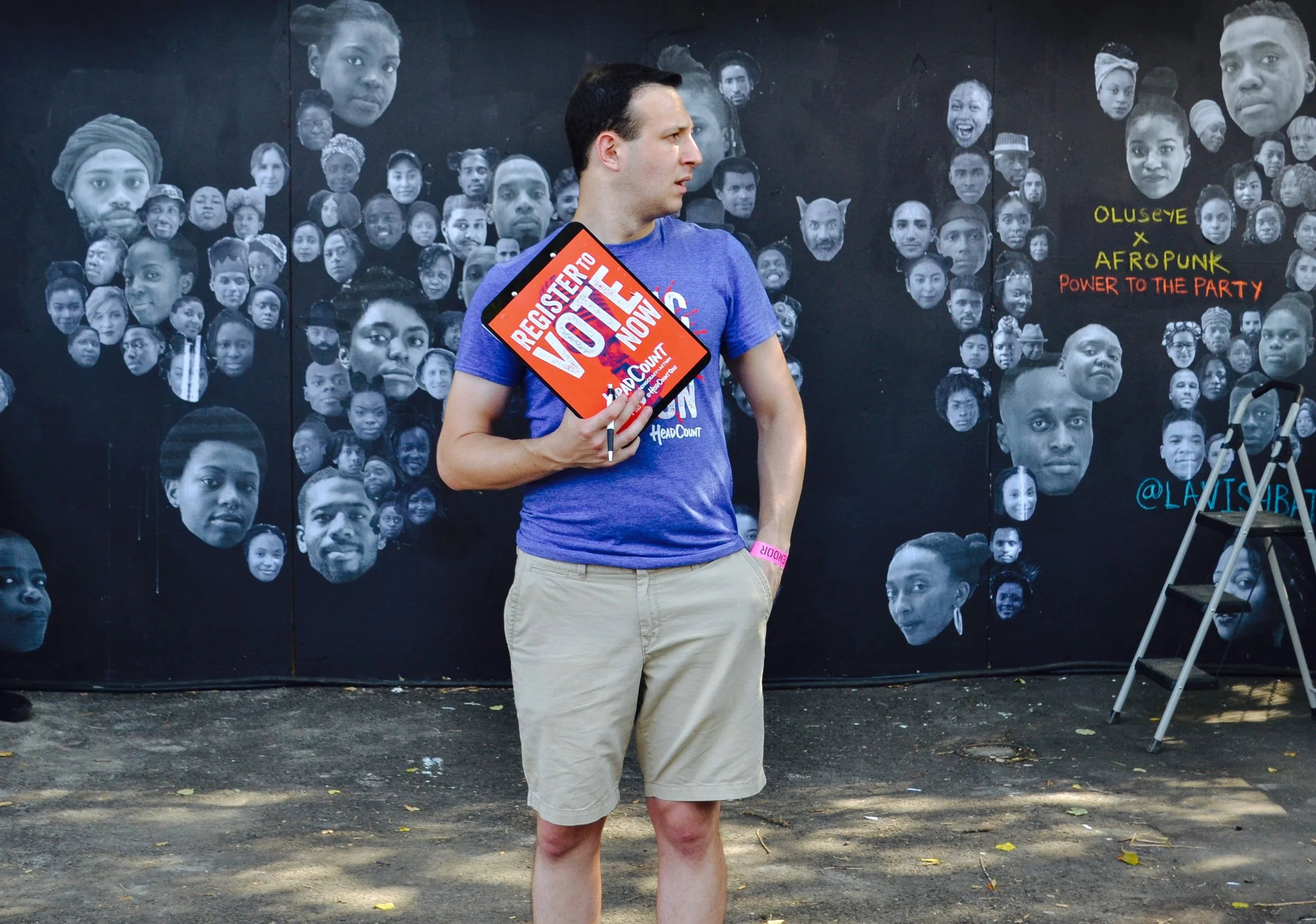 A man wearing a blue t-shirt and beige shorts standing outdoors in front of a black mural with numerous black-and-white photos of diverse faces. The man is holding a sign that reads 'Register to Vote Now' and has a pink wristband. There is a stepladd