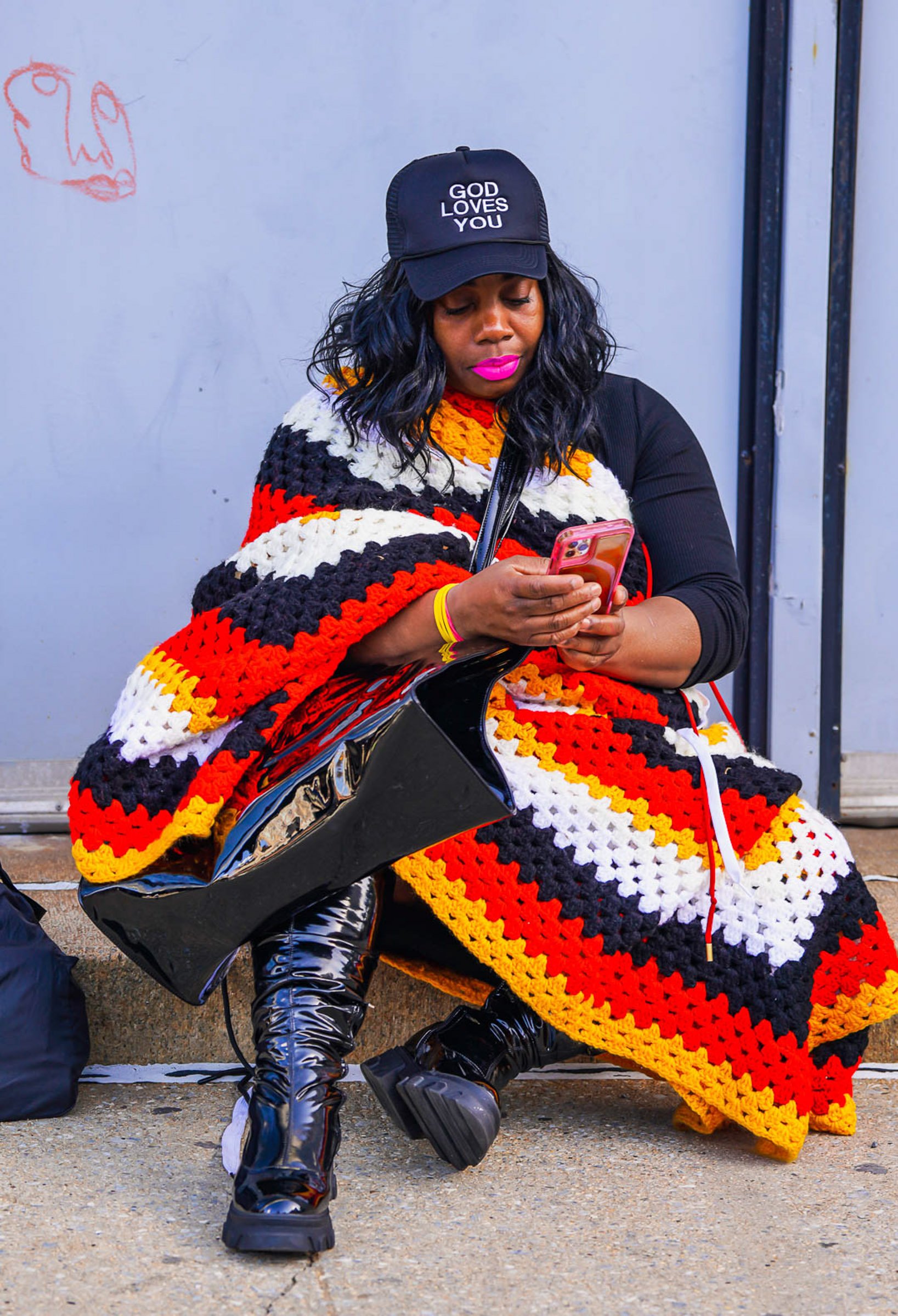 A woman with dark hair and pink lipstick sitting on a bench, wearing a black baseball cap that says 'God Loves You,' a black long-sleeve top, and shiny black pants. She is holding a pink phone and has a multicolored crochet blanket with red, yellow, 
